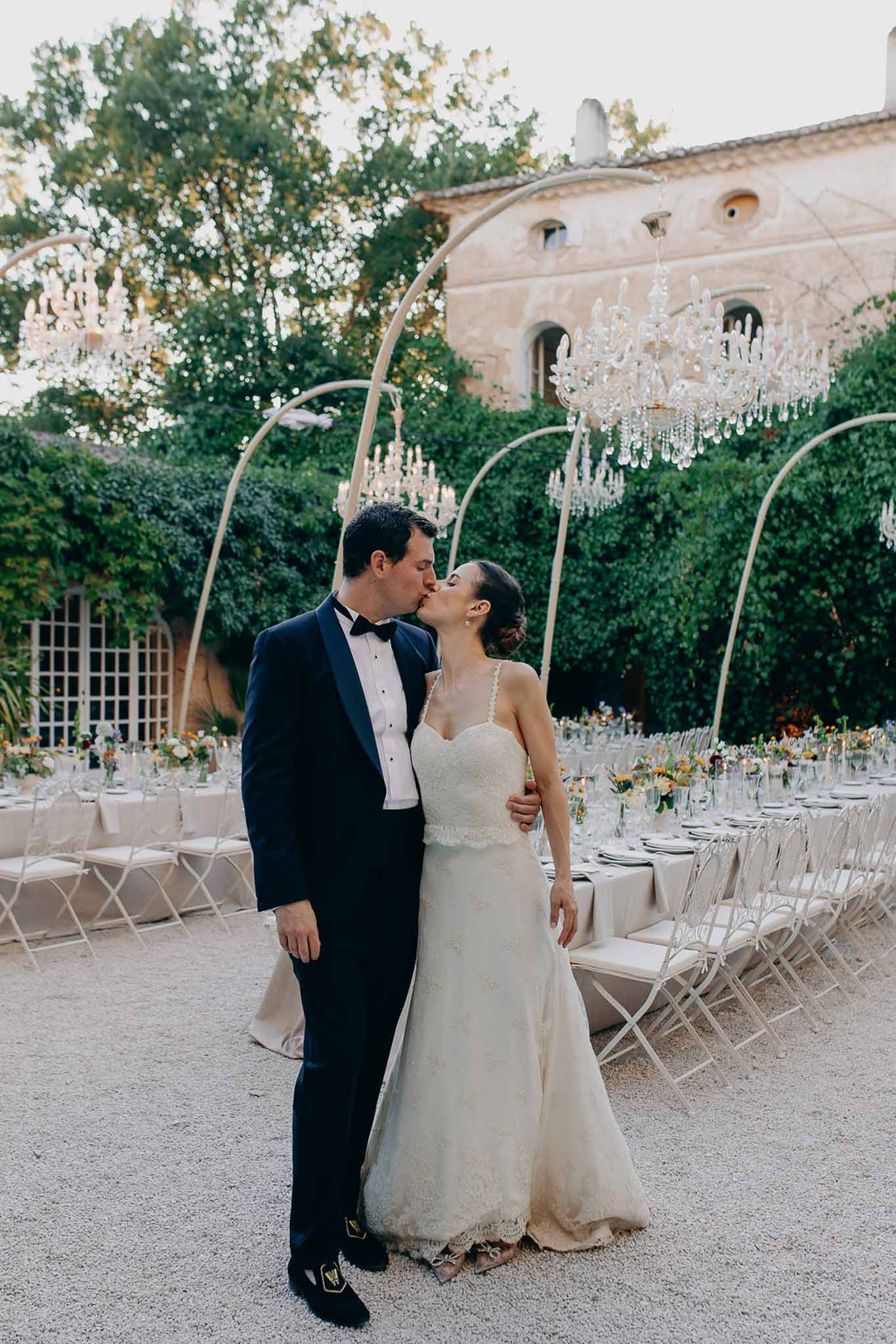 Couple kissing before courtyard reception with crystal chandeliers on gold arches banquet tables and ivy-covered stone walls