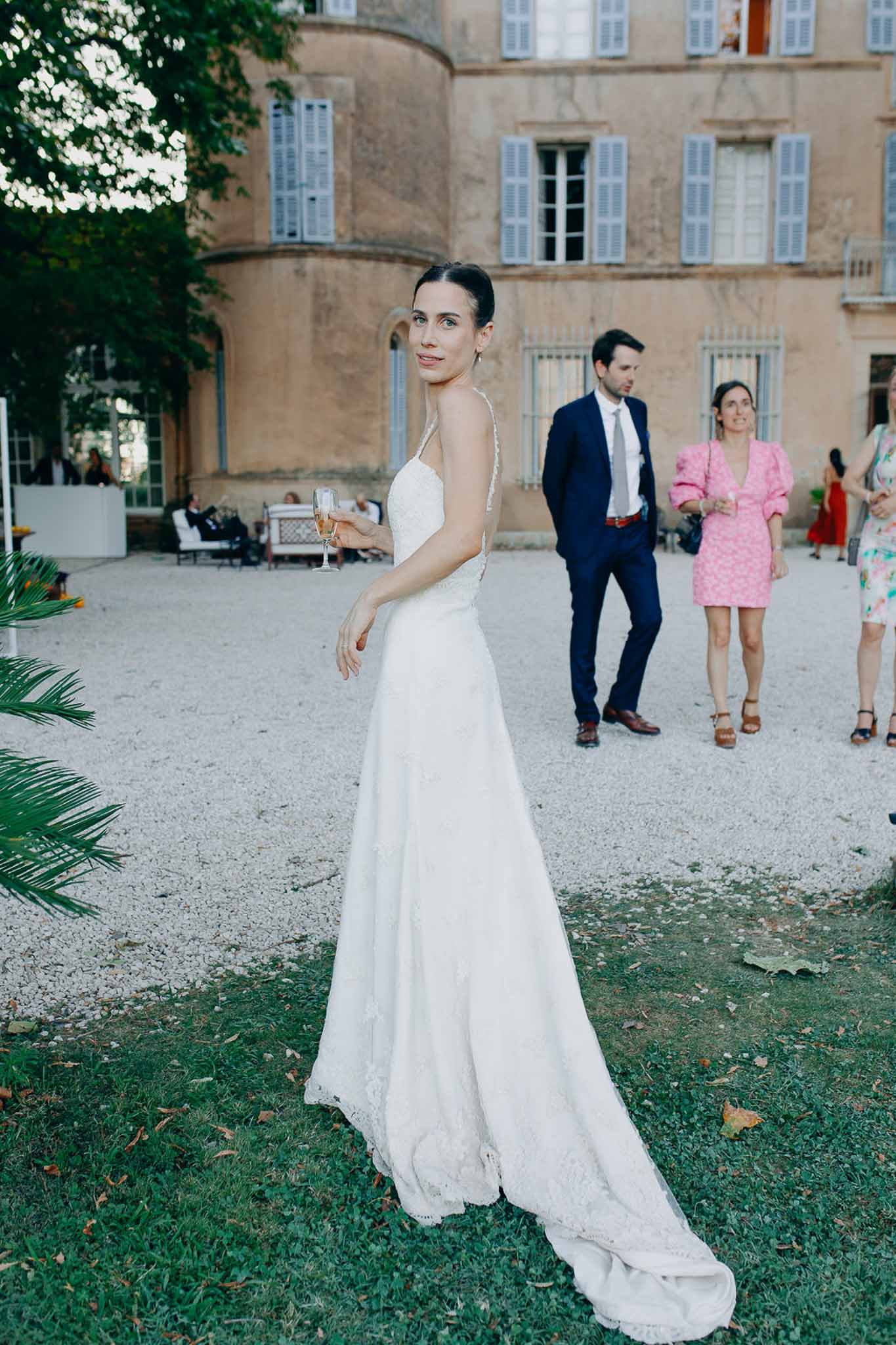 Bride in lace A-line gown holding rose champagne flute looking back at camera during cocktail hour