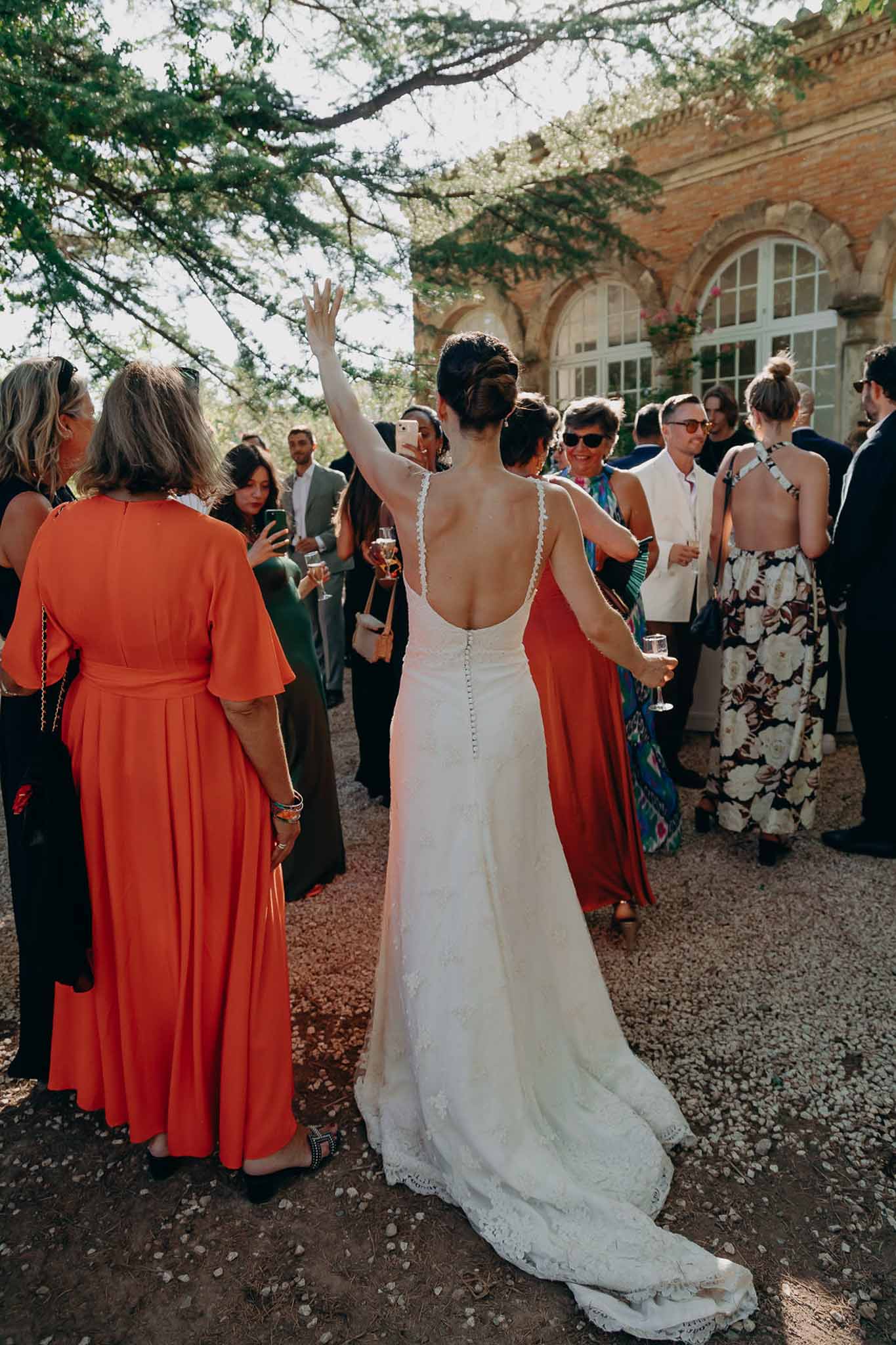 Bride raises champagne flute among 20 guests on gravel courtyard of red-brick chateau from behind