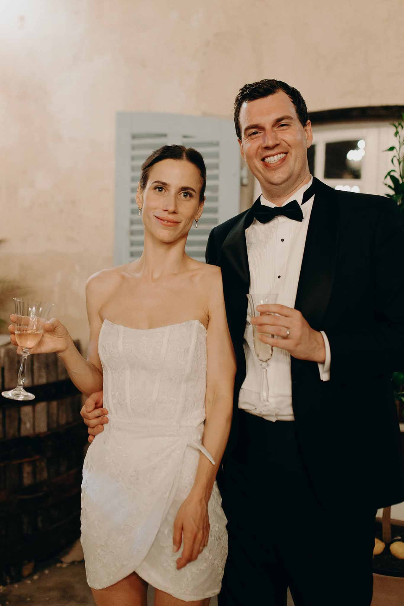 Couple holding rose champagne bride in white embroidered mini dress groom in tuxedo by rustic shutter and barrel