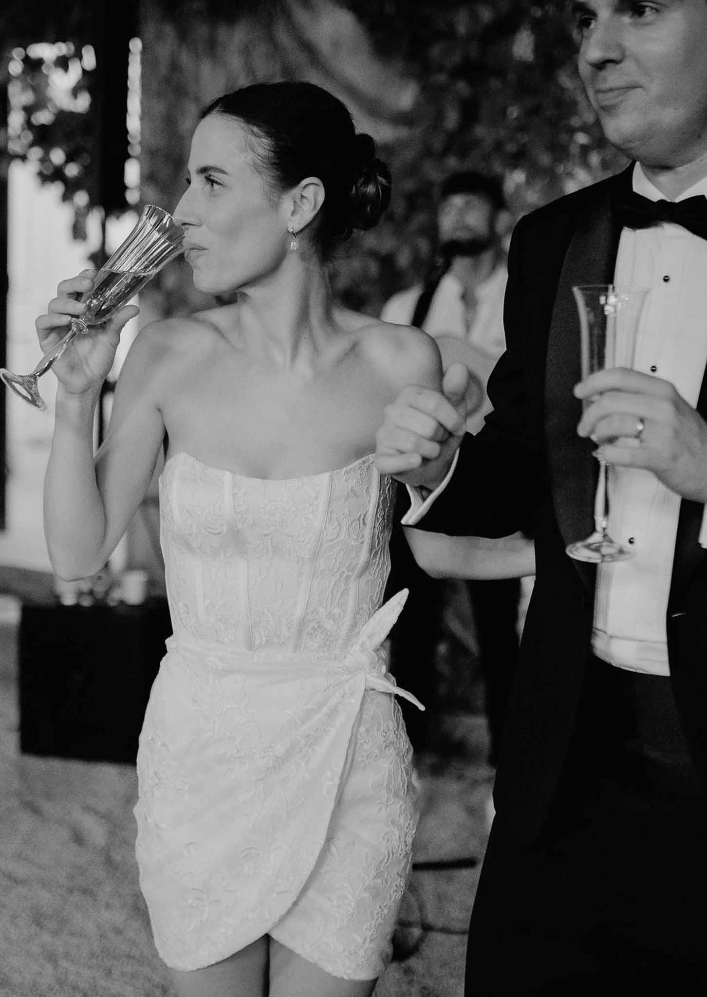Black and white bride in short strapless lace dress sipping champagne with groom in tuxedo at reception