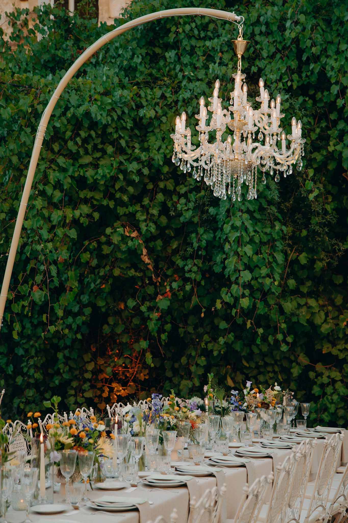 Garden table with wildflower arrangements crystal chandelier on ivory arch taper candles and iron chairs