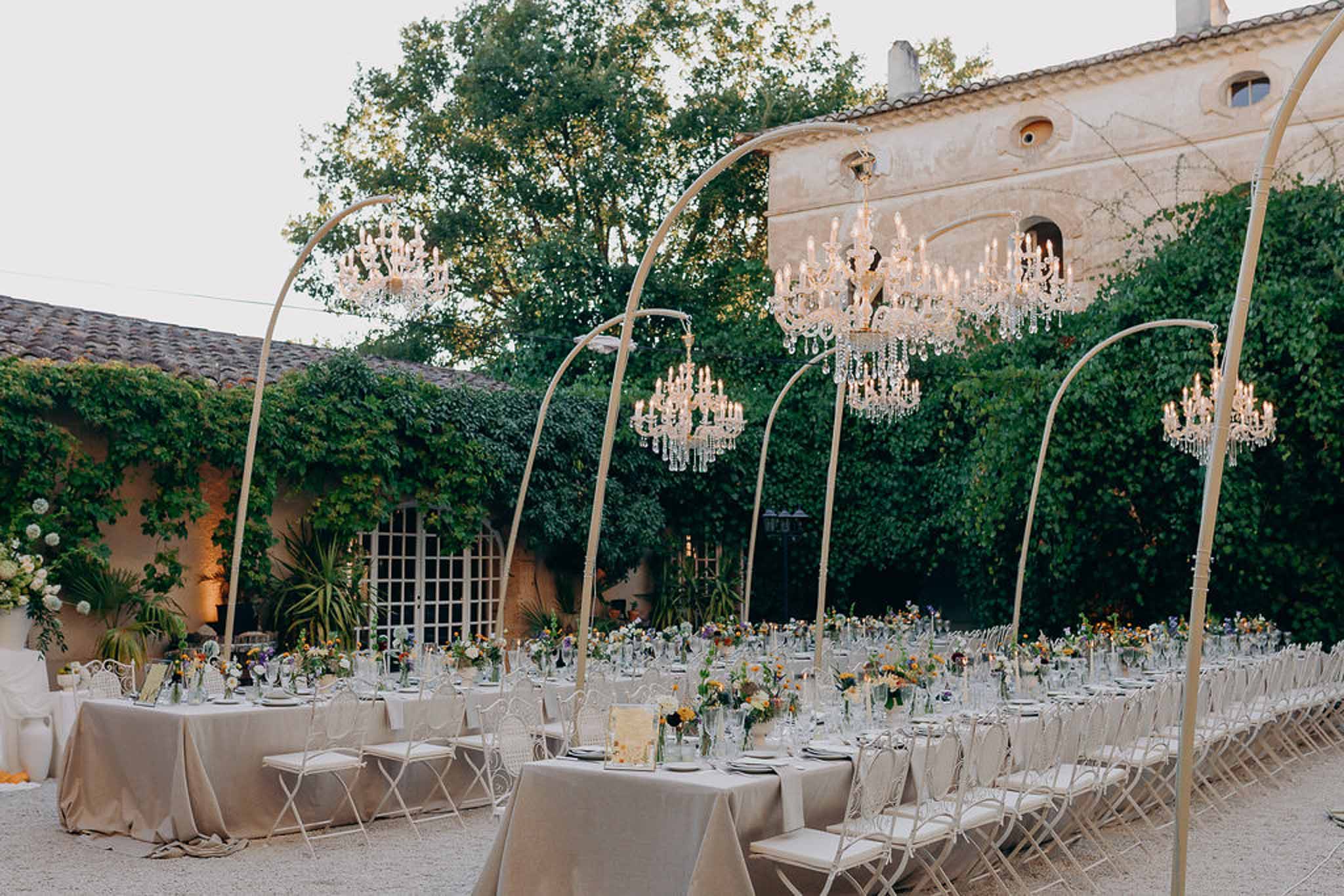 Dusk courtyard reception with crystal chandeliers on gold arching poles taupe linen tables and colorful bud vase centerpieces