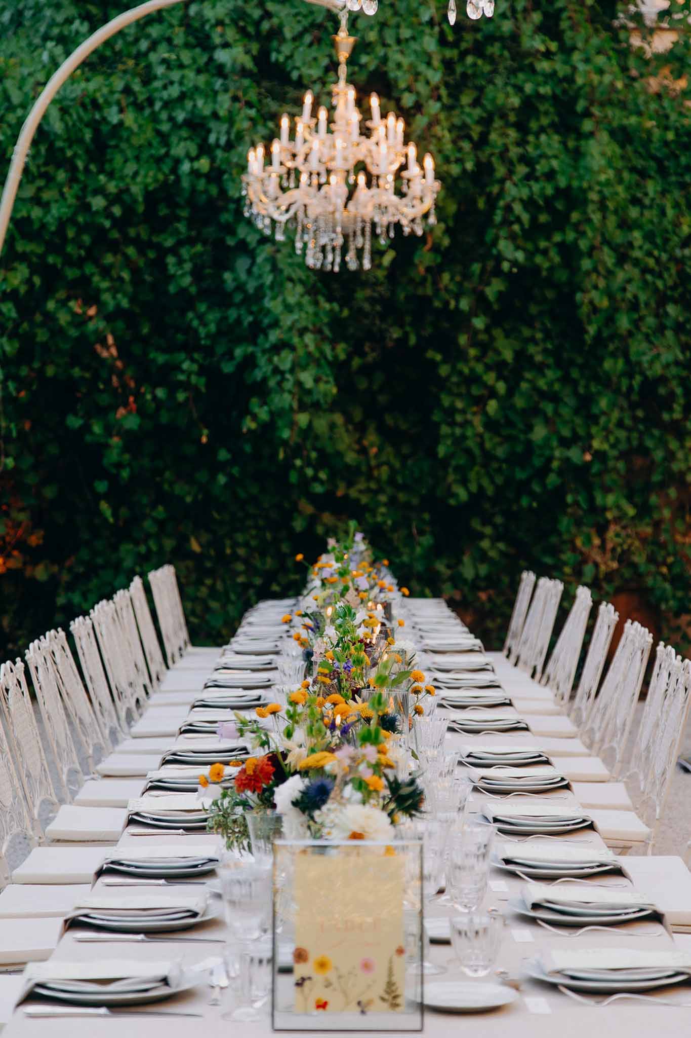 Long outdoor reception table with wildflower runner of yellow and orange blooms set against an ivy-covered wall with cryst...