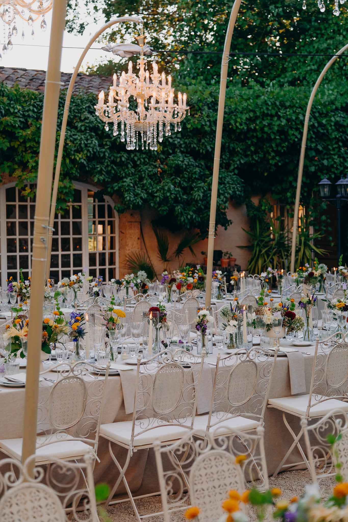 Dusk courtyard reception with crystal chandeliers on arched frames colourful wildflower centerpieces and iron chairs