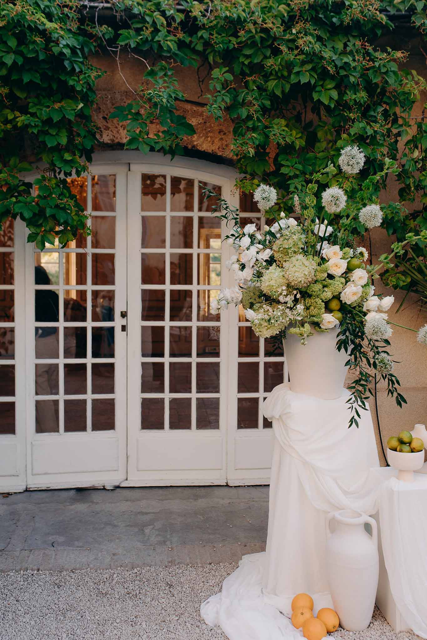 White cake beside ceramic urn of green hydrangeas and white roses with limes at vine-framed doors