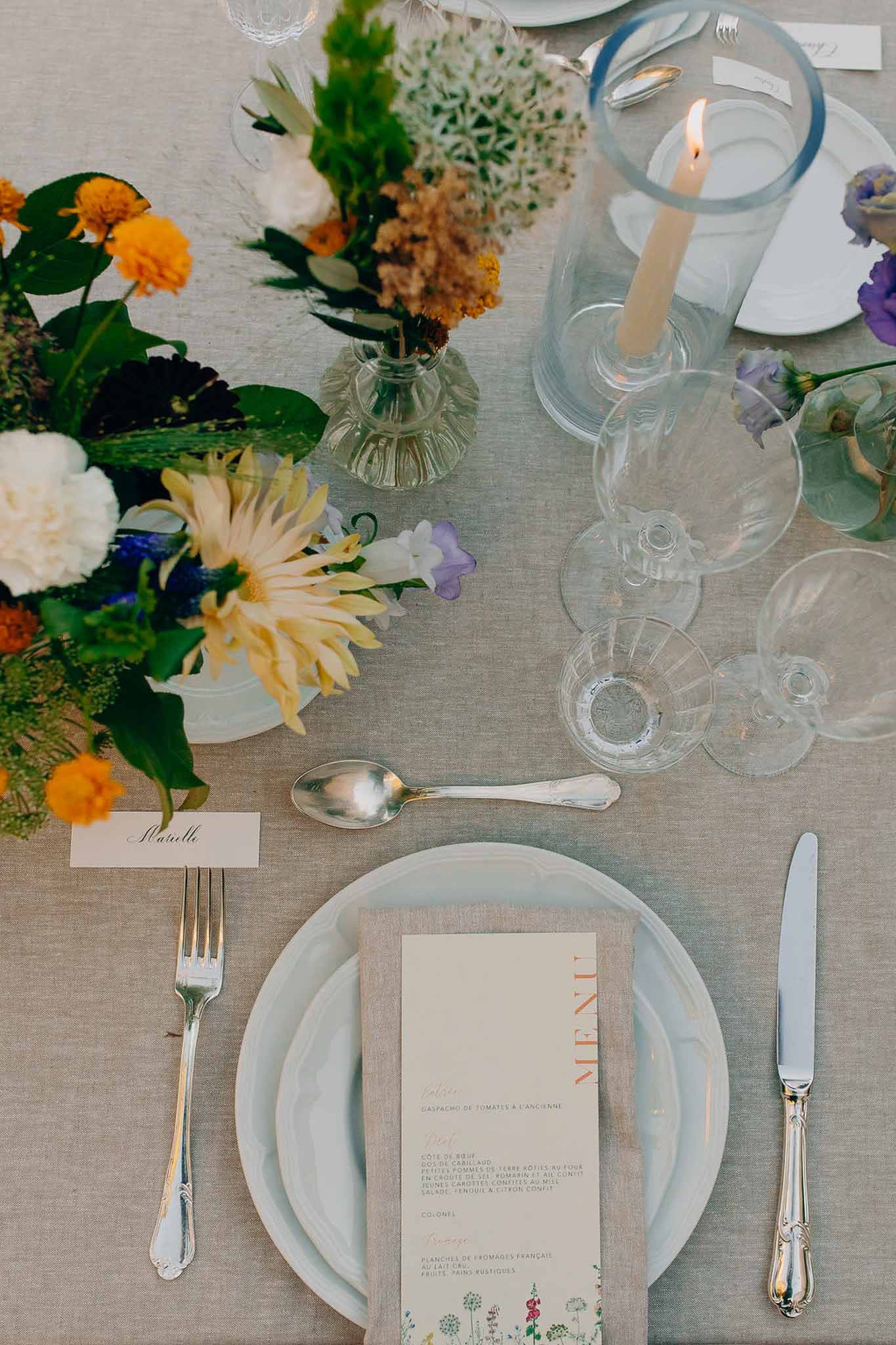 Place setting with coral-lettered French menu, marigold and blue bud vases, and ivory taper candle
