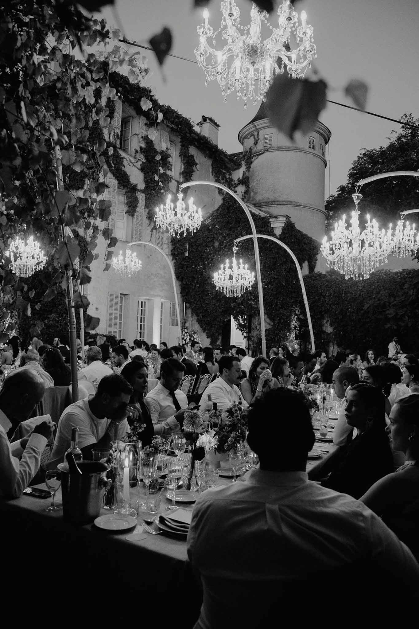 Outdoor chateau courtyard reception at dusk with crystal chandeliers and guests at long tables in black and white