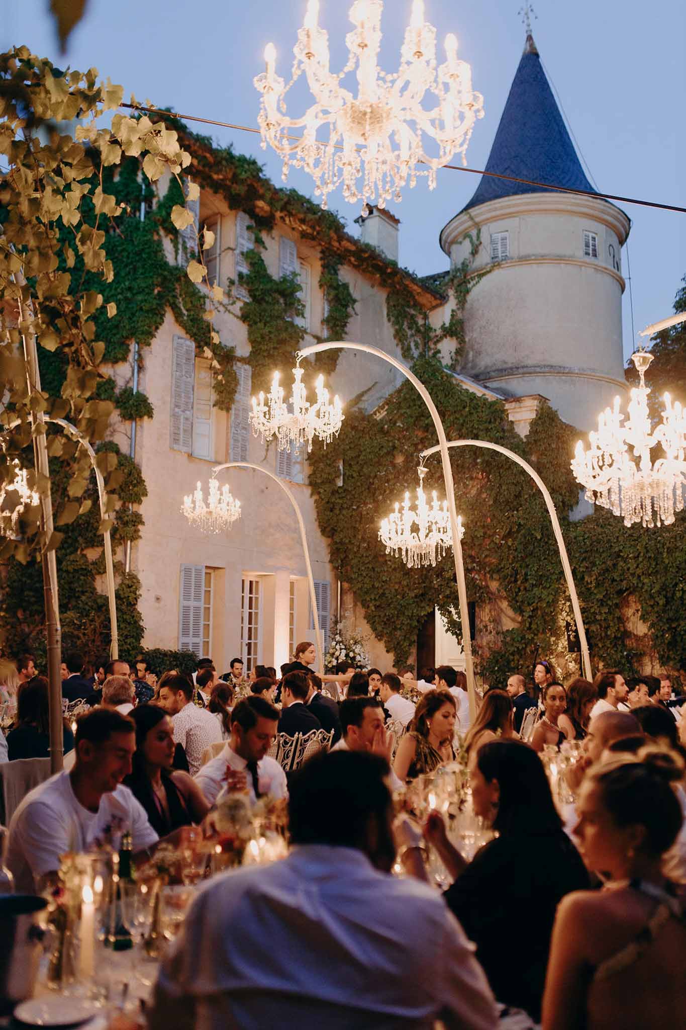 Outdoor evening reception in an ivy-covered chateau courtyard with guests seated at long tables under suspended crystal ch...