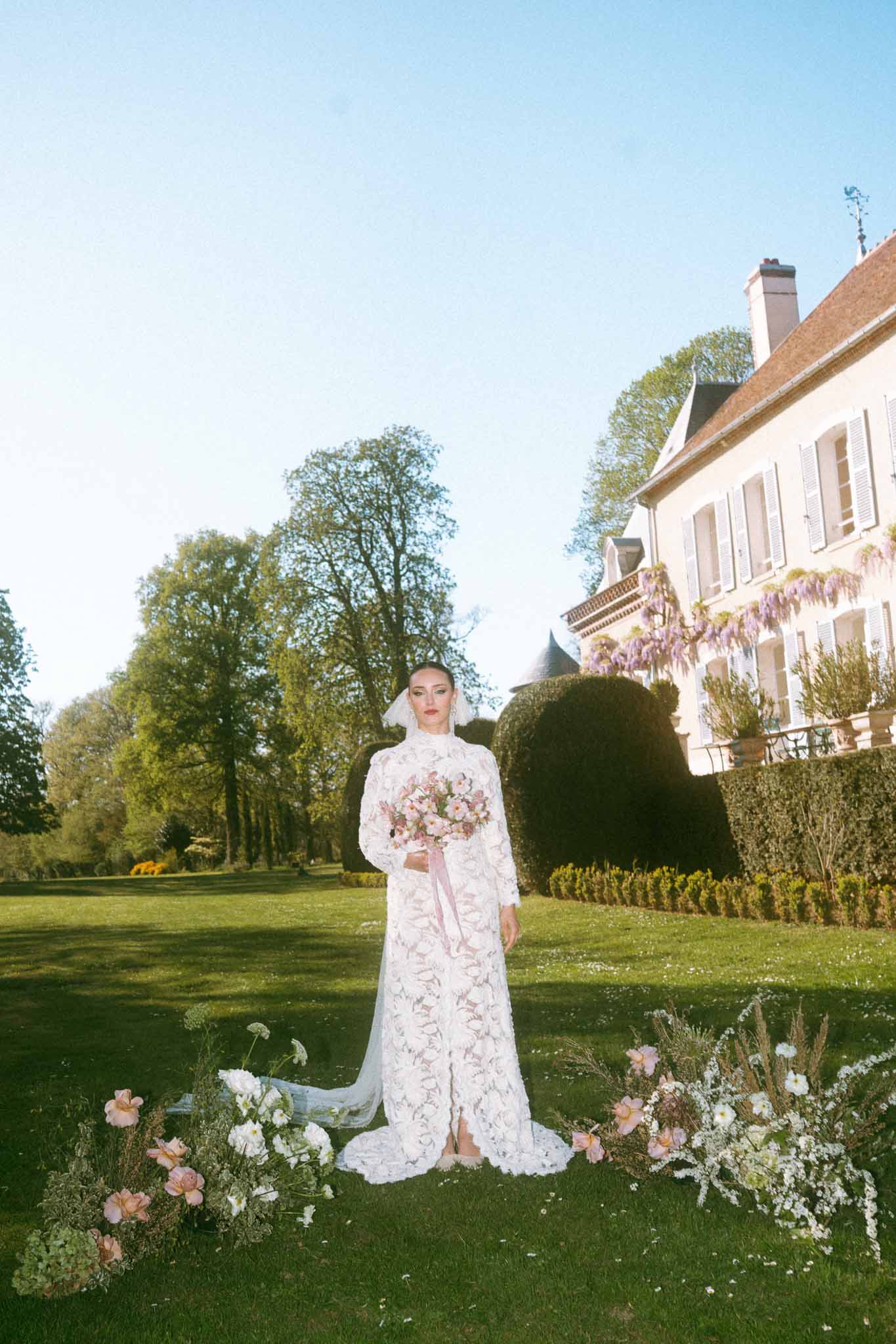 Bride in ivory lace gown with blush bouquet standing on formal estate garden lawn