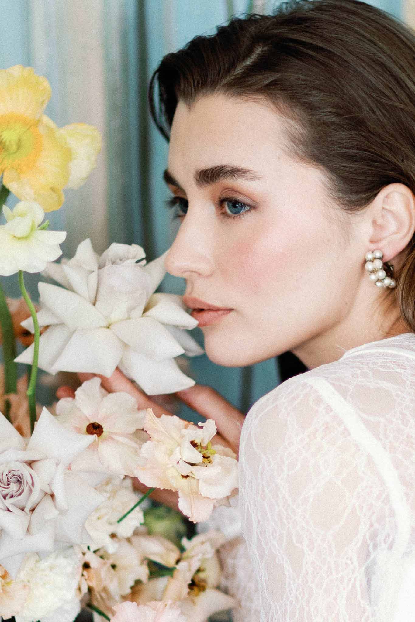 Bride in white lace dress holding bouquet of orchids, anemones, and lavender roses up to her face