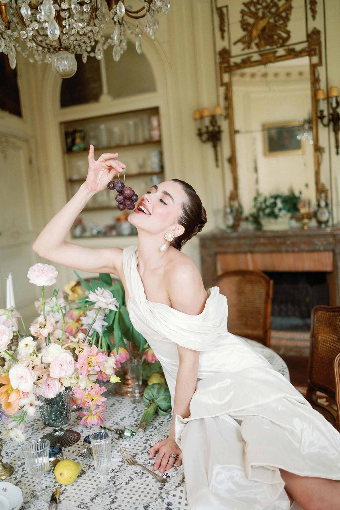 Bride in one-shoulder gown with grapes at editorial table with coral ranunculus in gilded chateau salon