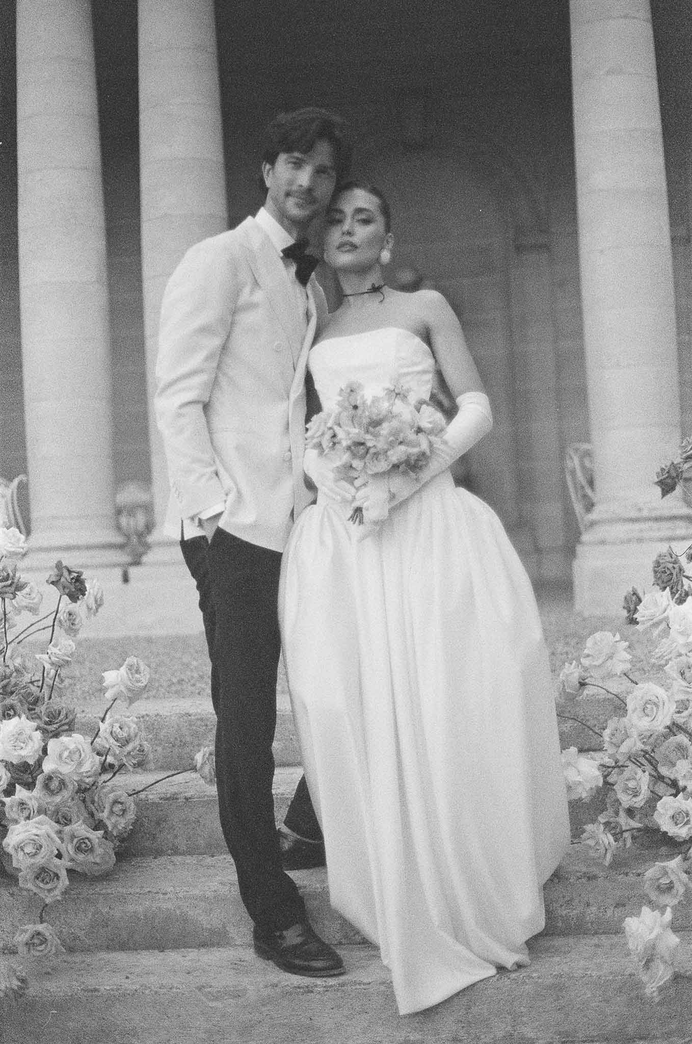 Black and white of couple on columned chateau steps with rose arrangements, bride in gloves and choker