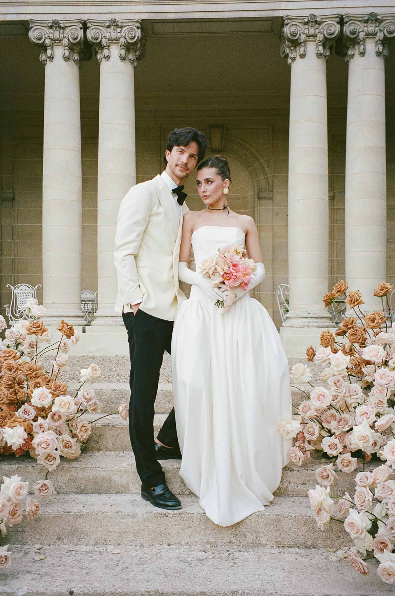 Couple on neoclassical steps flanked by blush and peach rose clusters, bride in ballgown with gloves and pink bouquet