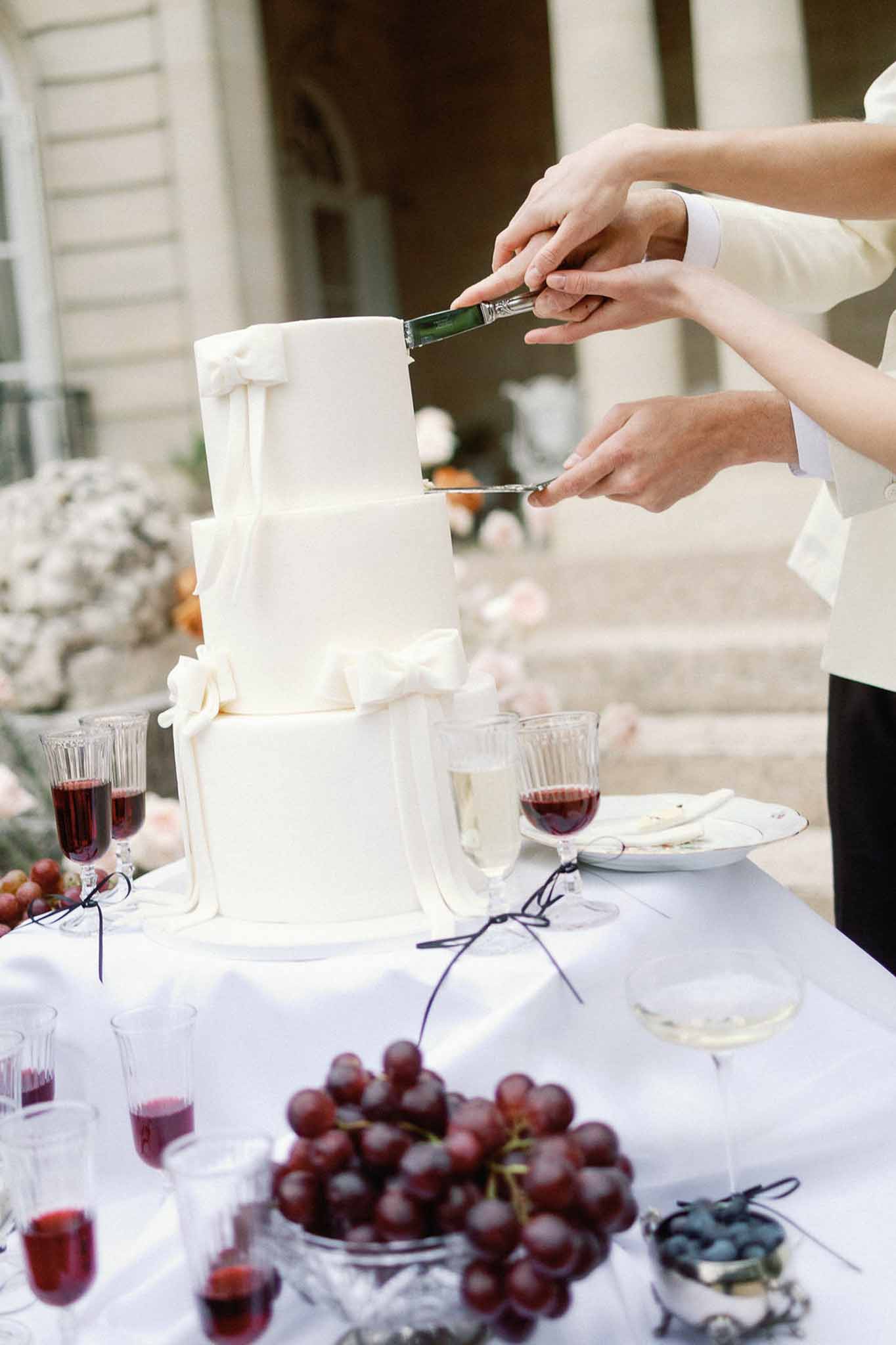 Couple cutting three-tiered ivory fondant wedding cake on table styled with red grapes and crystal glassware