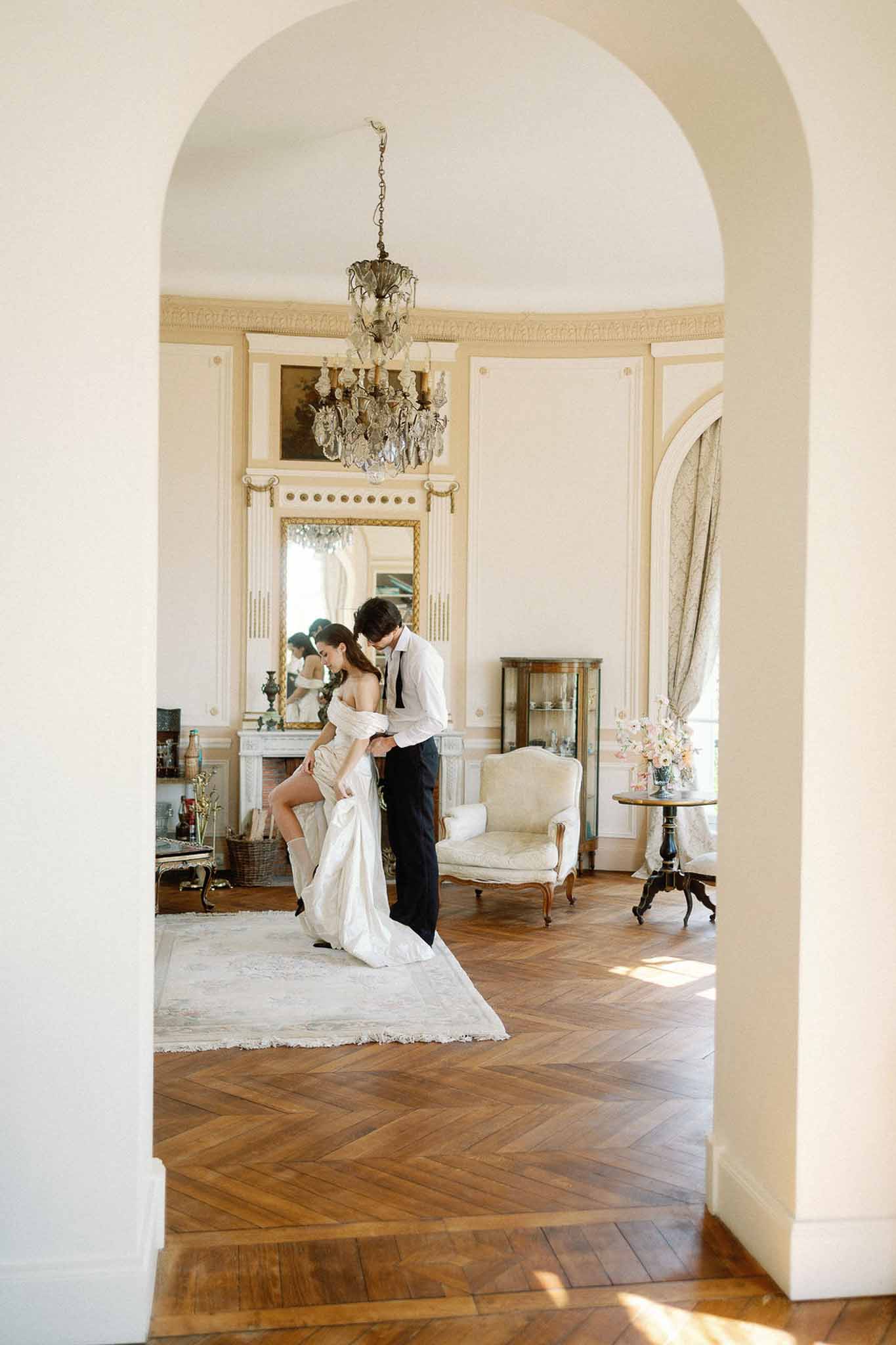 Groom helps bride with dress in ornate chateau room with crystal chandelier and herringbone parquet floor