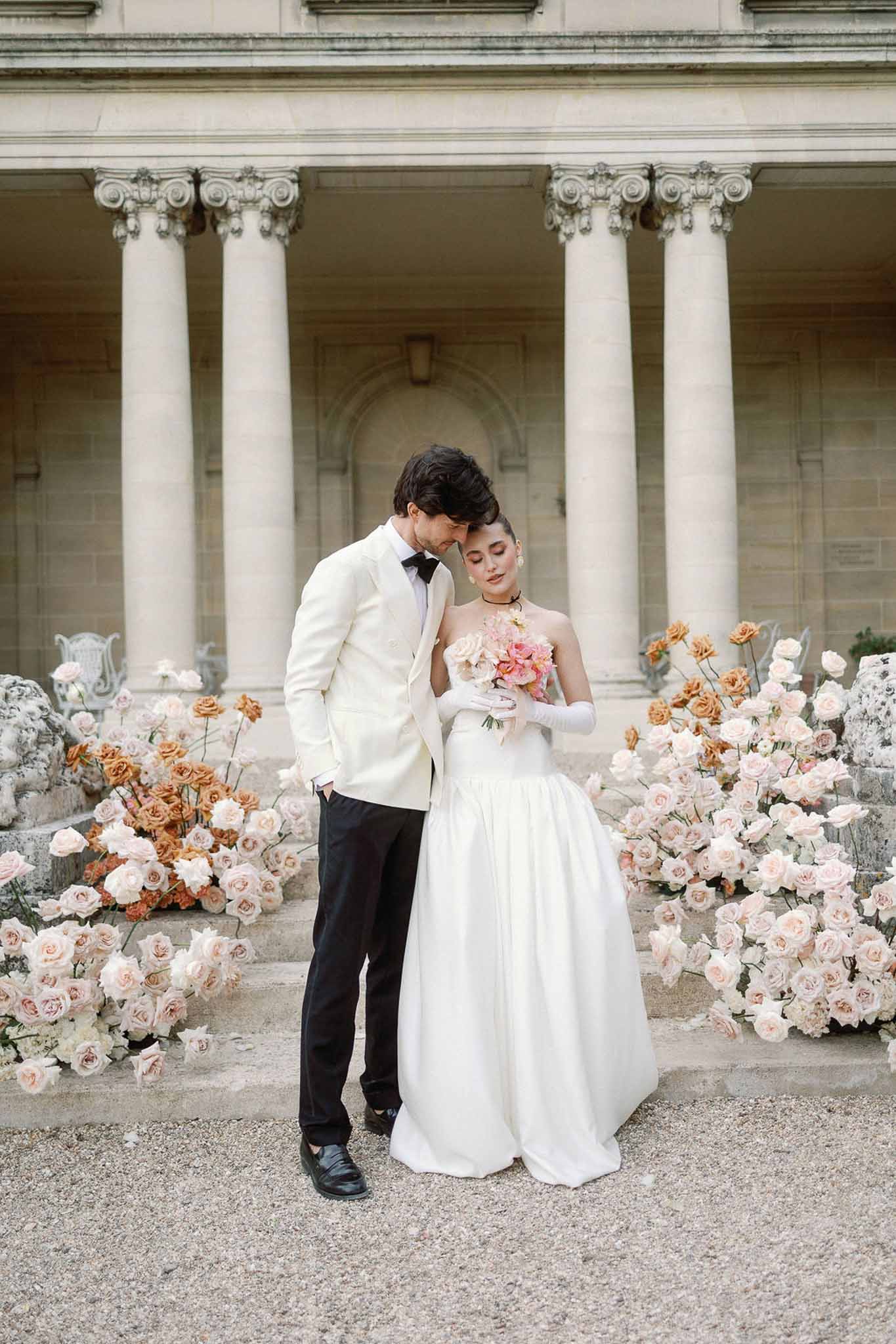 Bride in white ballgown and groom in ivory dinner jacket touching foreheads on neoclassical steps with pink rose arrangements