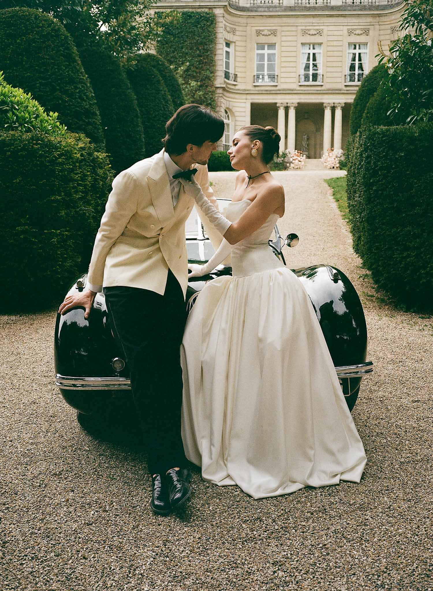 Bride in ivory ball gown and groom in ivory dinner jacket posed on a vintage black car at a neoclassical chateau