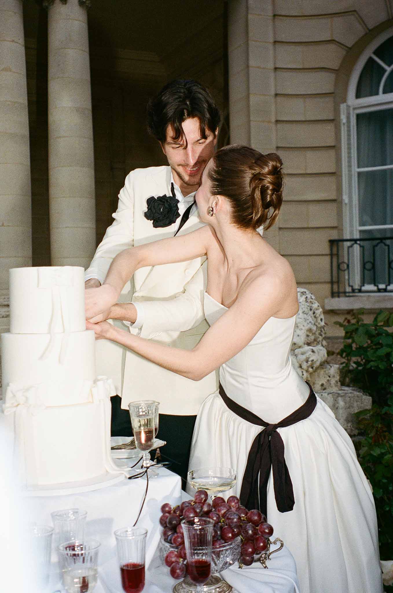 Couple cutting four-tier white fondant cake with grapes and crystal glassware at chateau facade