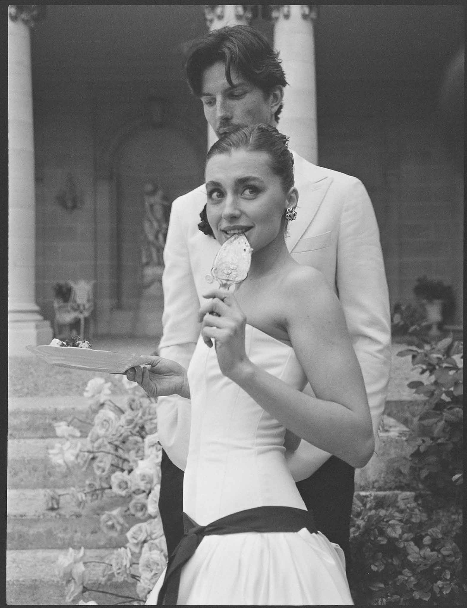 Black and white portrait of bride eating cake while groom stands behind her on chateau steps lined with garden roses