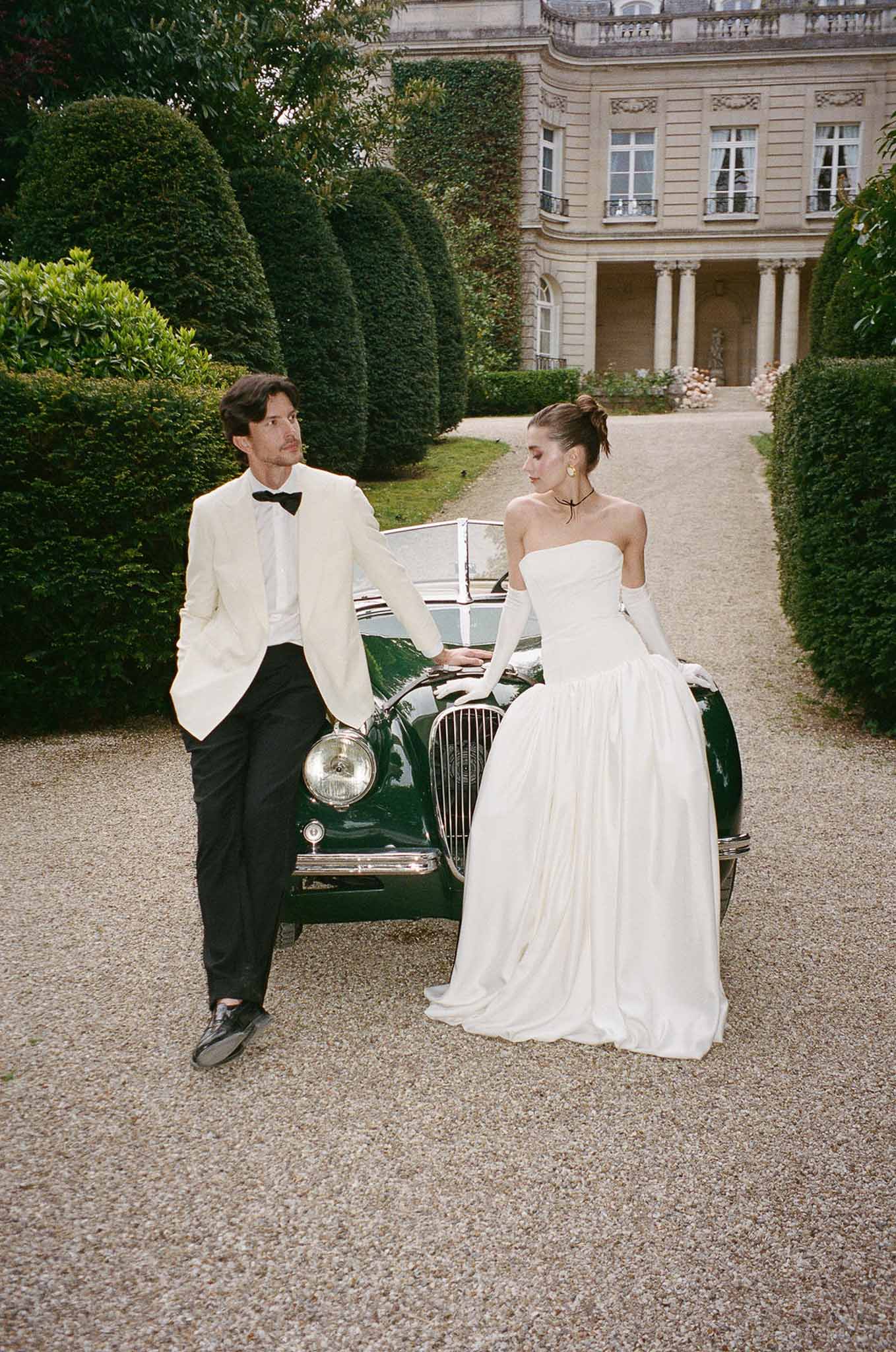 Couple in ivory dinner jacket and ballgown with gloves leaning on vintage green Jaguar at chateau drive
