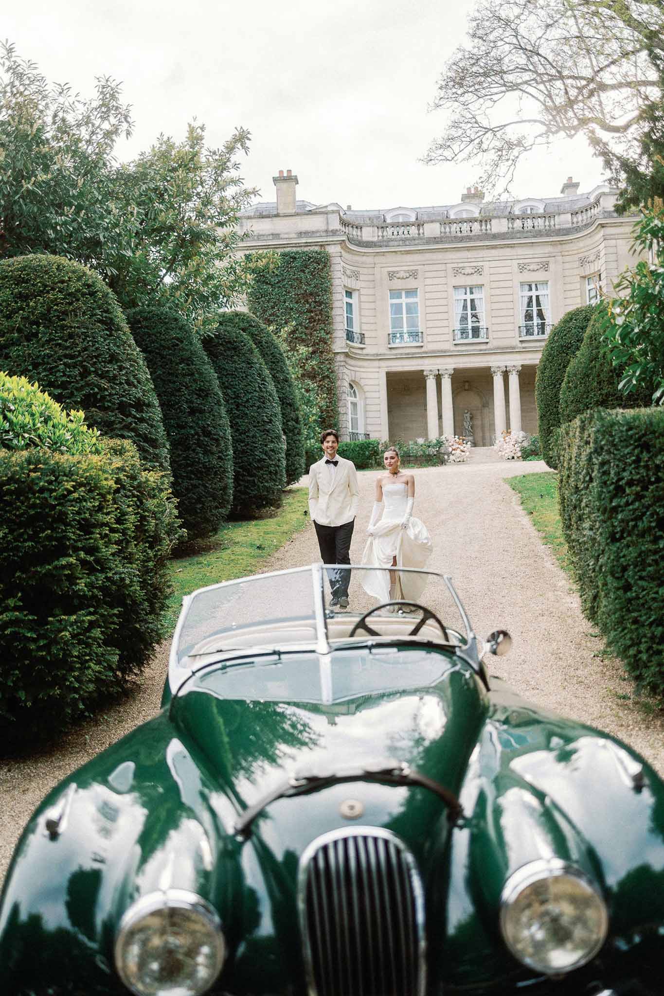Bride and groom walking on chateau driveway past a vintage green convertible with neoclassical facade behind
