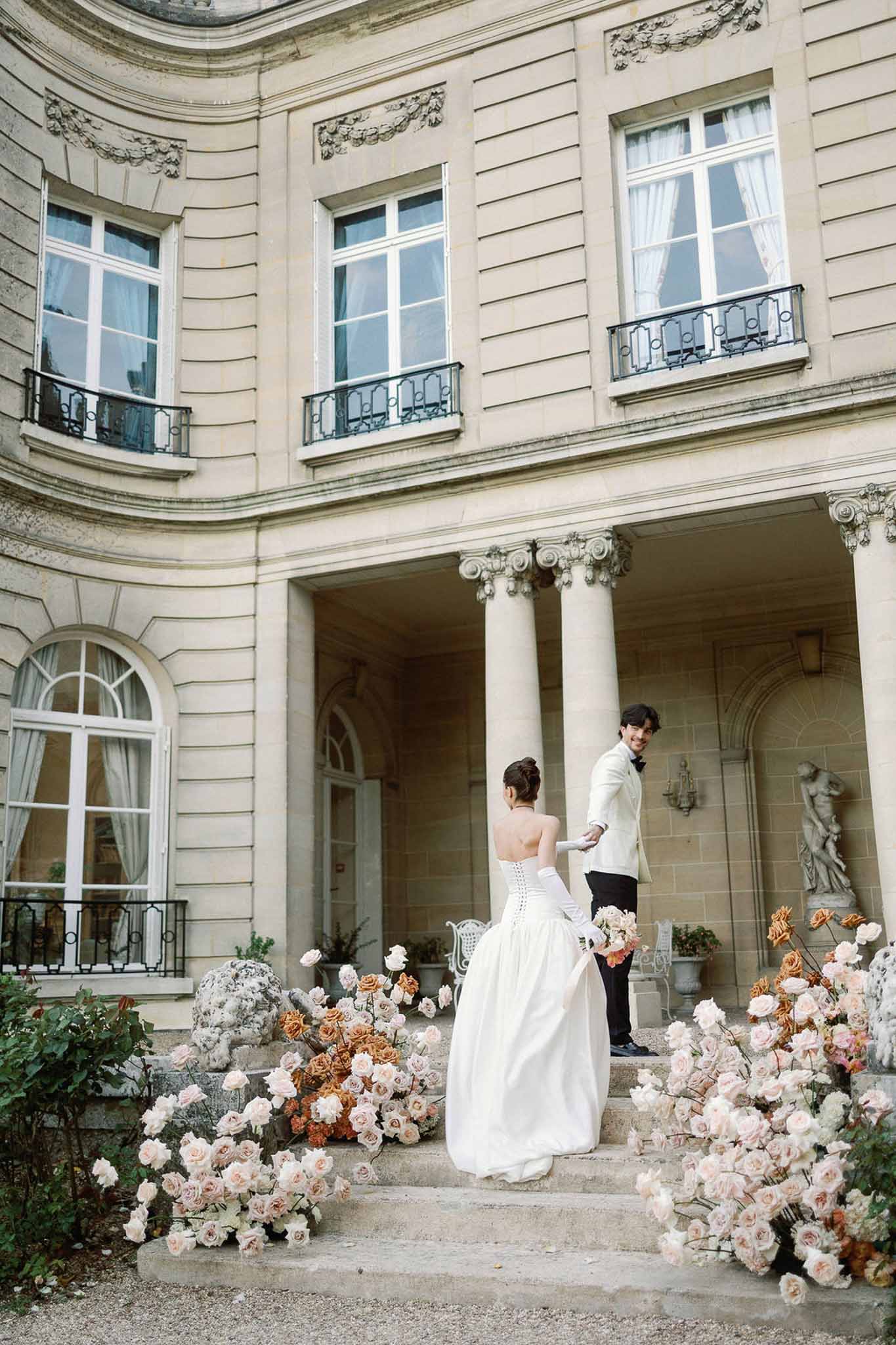 Bride and groom on steps of classical mansion with blush and burnt orange rose-lined staircase