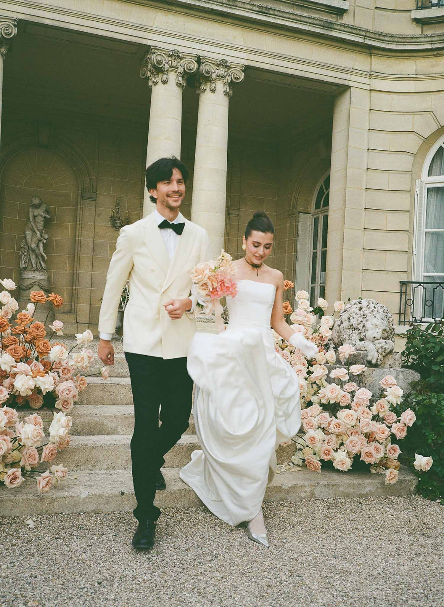 Couple descends chateau stairs lined with coral roses as bride carries matching bouquet in film tones