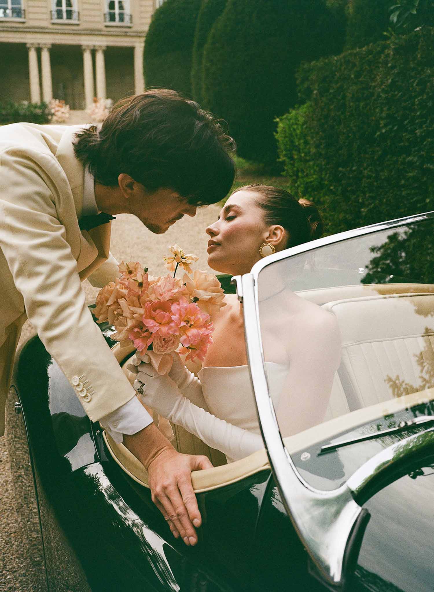 Groom leaning over vintage green convertible to kiss bride holding coral peony bouquet