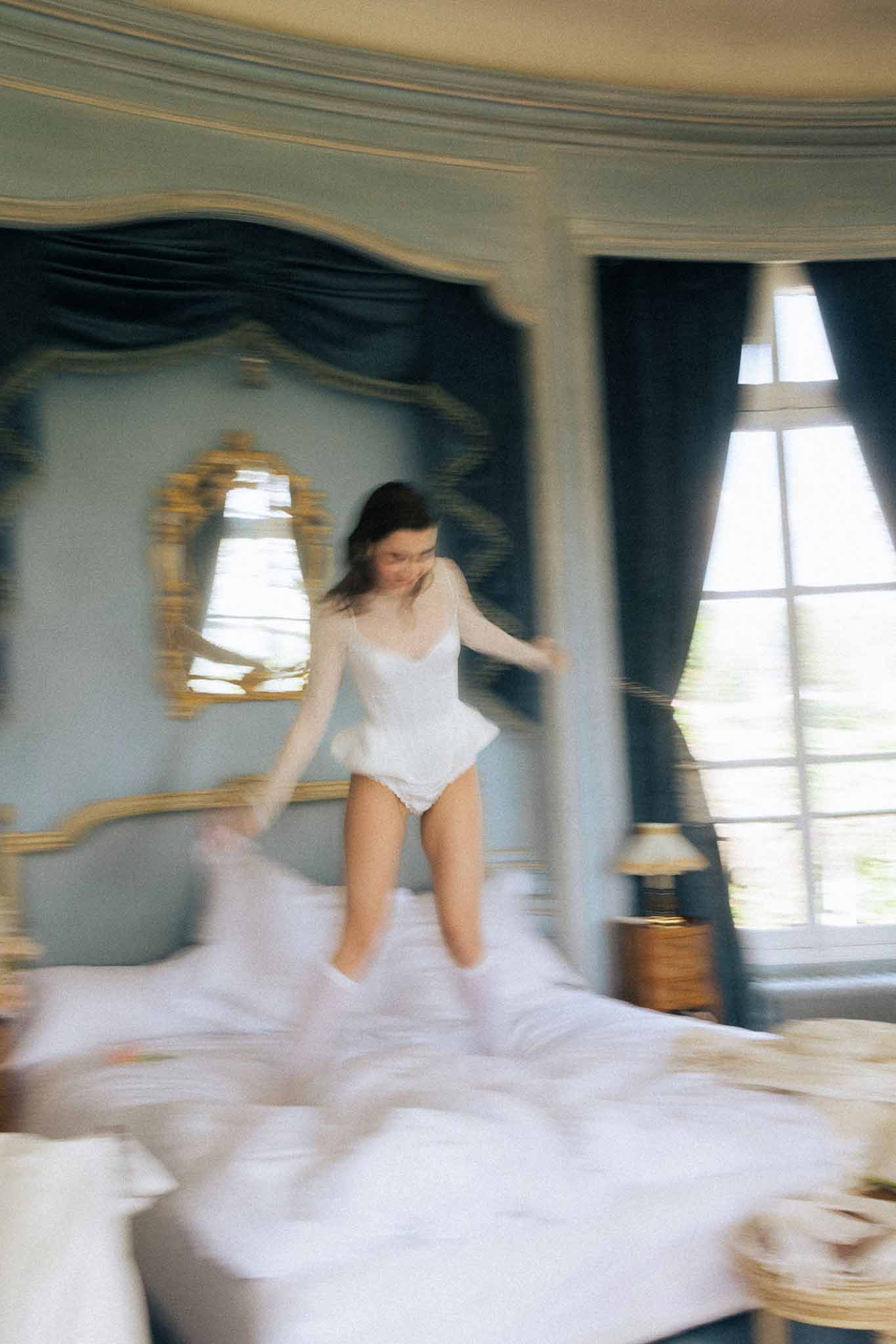 Bride in white lace bodysuit jumping on bed in blue-panelled chateau bedroom with gold moulding