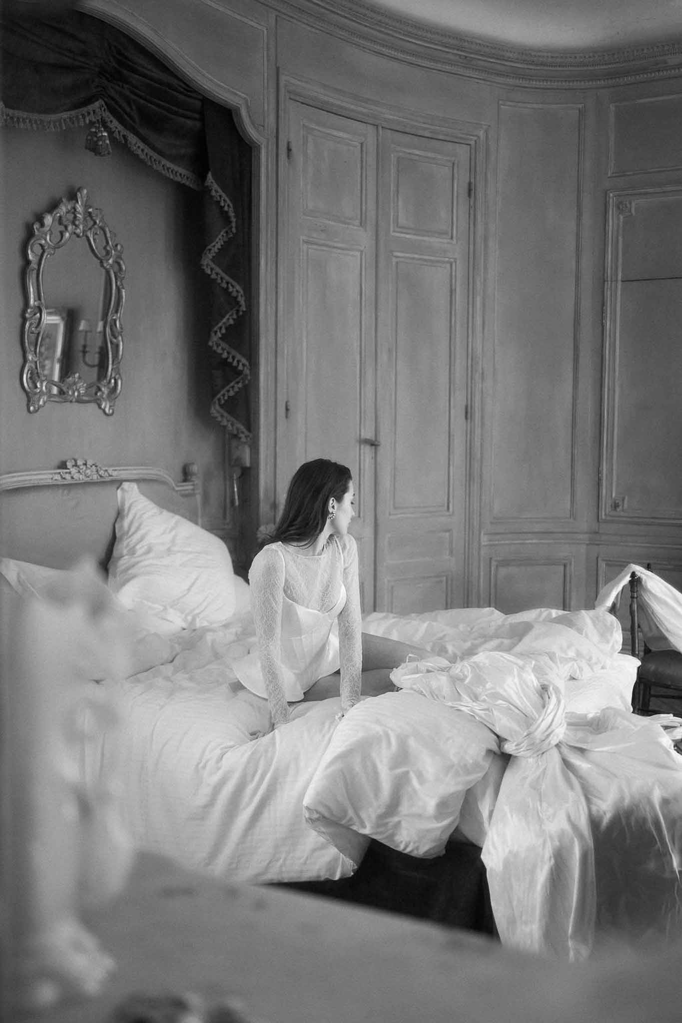Black and white of bride in lace top seated on chateau bed beside laid-out gown with bow detail and gilt mirror