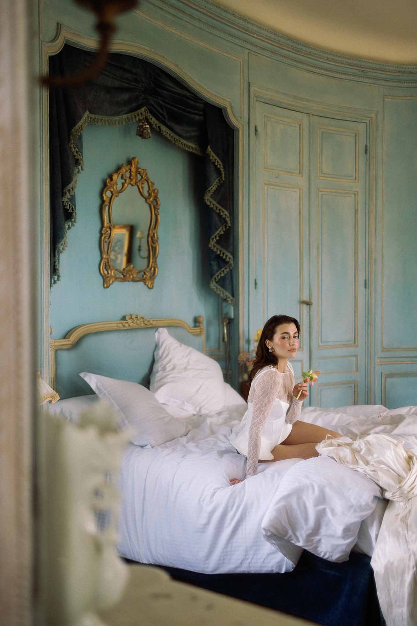 Bride in white lace top seated on bed in duck-egg blue panelled chateau room with gilded mirror and teal canopy