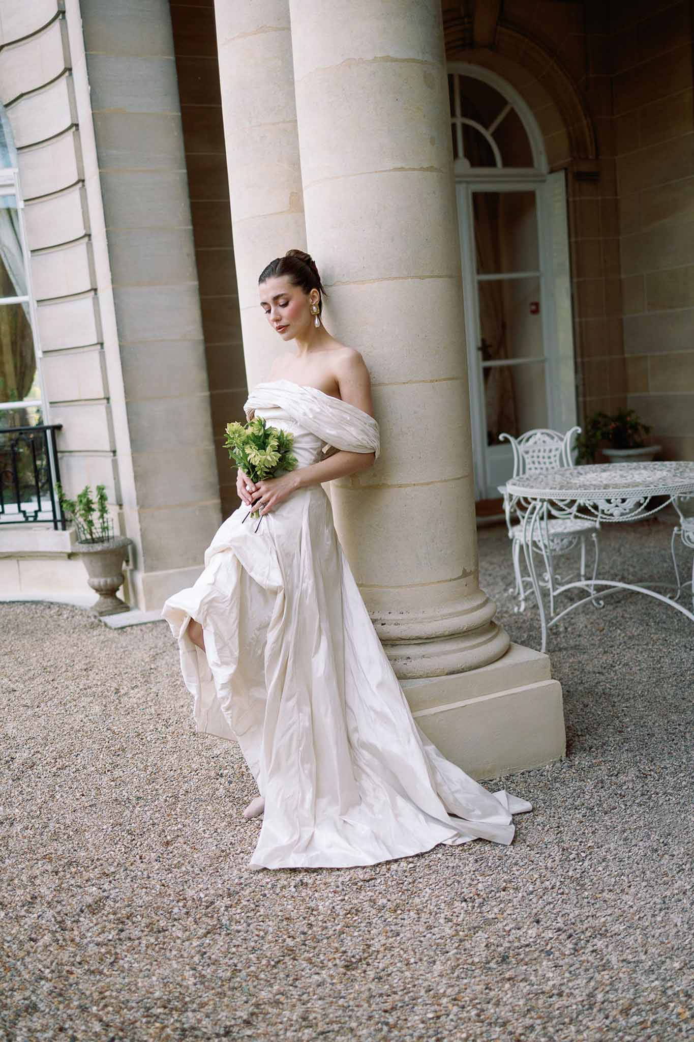 Bride in off-the-shoulder ivory ball gown leaning against stone column holding green bouquet at French chateau forecourt