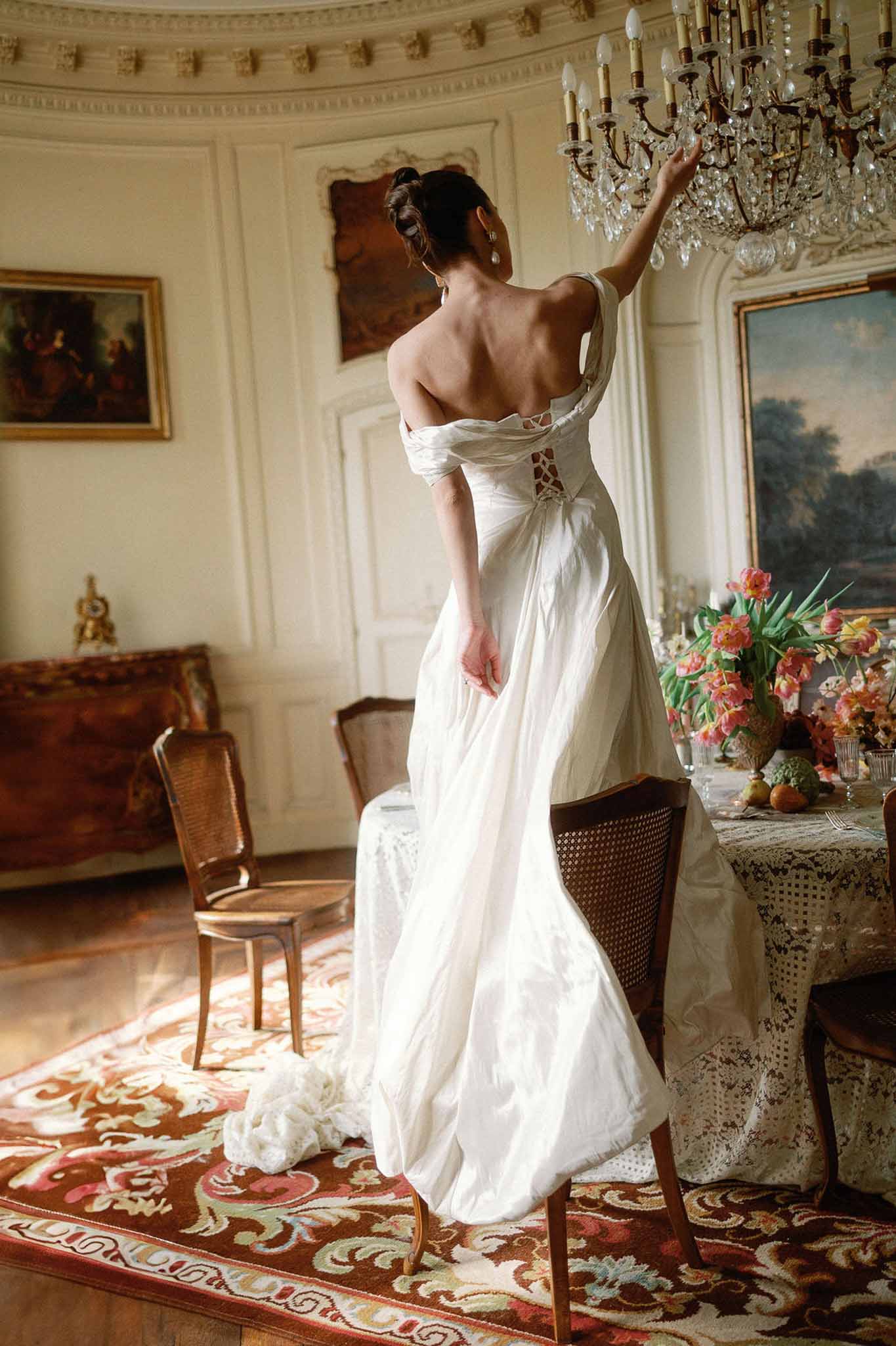 Bride in off-the-shoulder ivory gown reaching toward chandelier in ornate chateau salon with gilded walls and tulip arrang...
