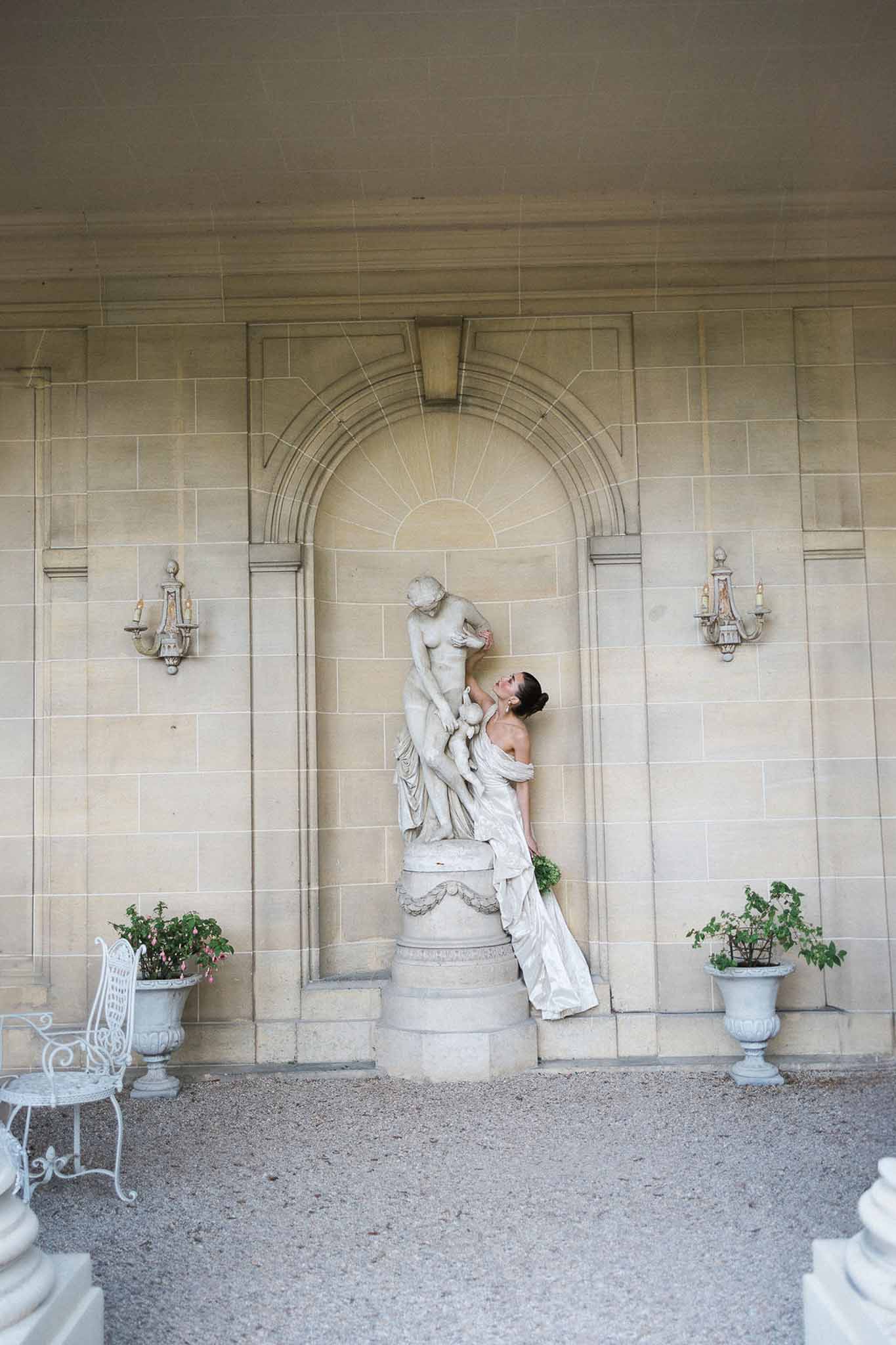 Bride leaning against marble statue in stone niche at chateau portico with urn planters and iron sconces