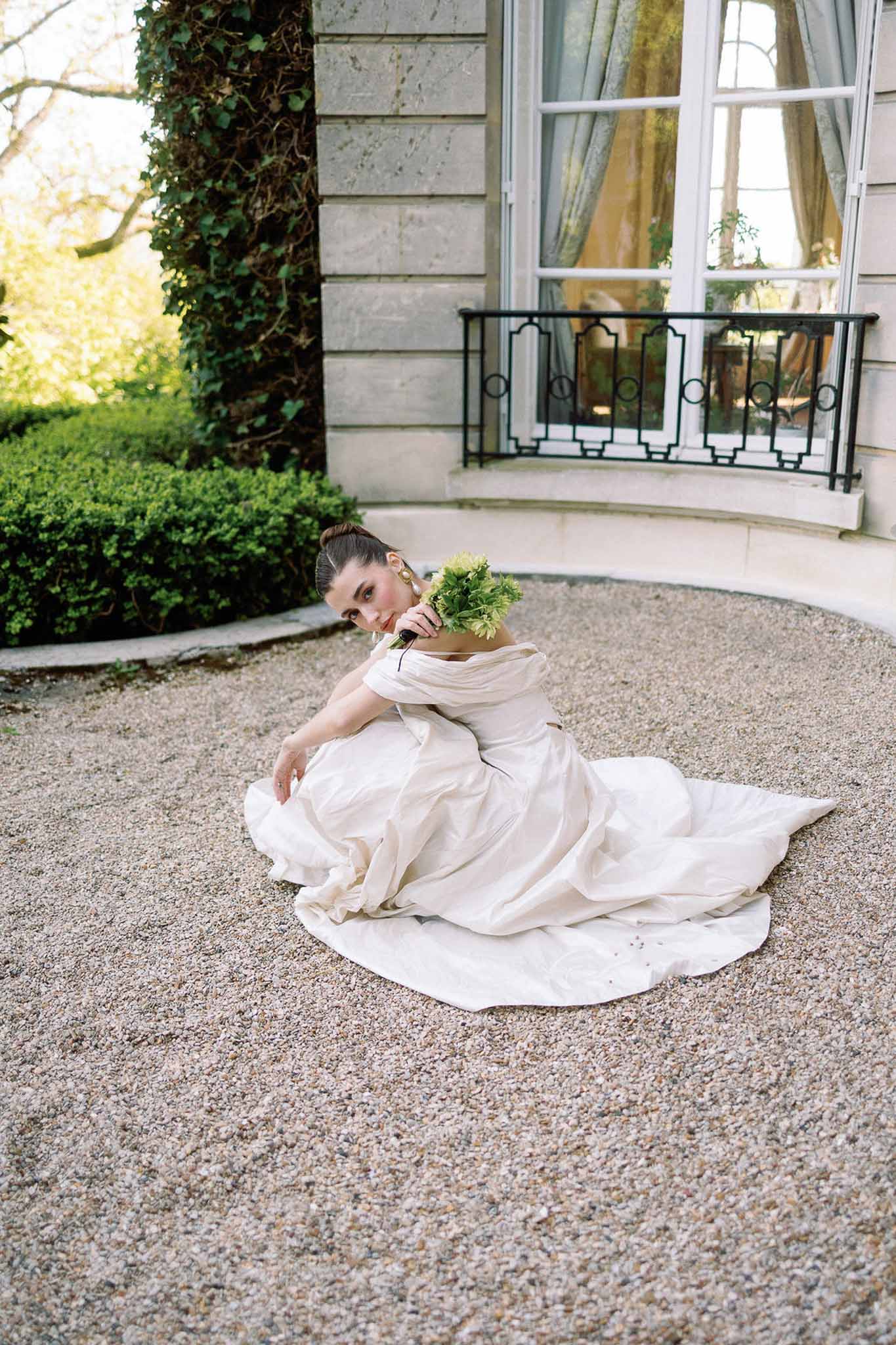 Bride seated on gravel with ivory off-shoulder gown spread wide holding chartreuse bouquet before chateau