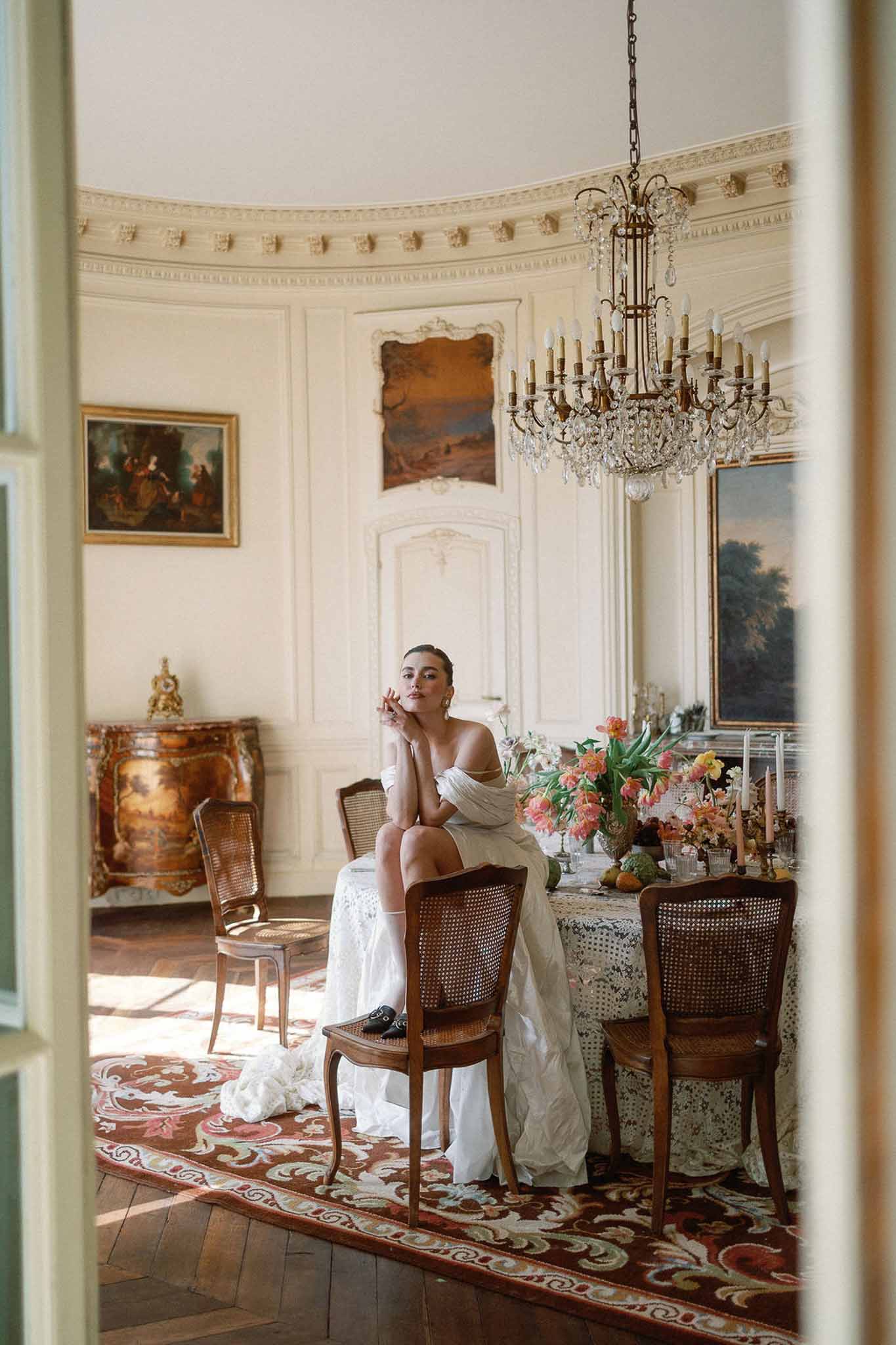 Bride in off-the-shoulder ivory gown seated on a dining table in a panelled chateau room with coral tulips and chandelier