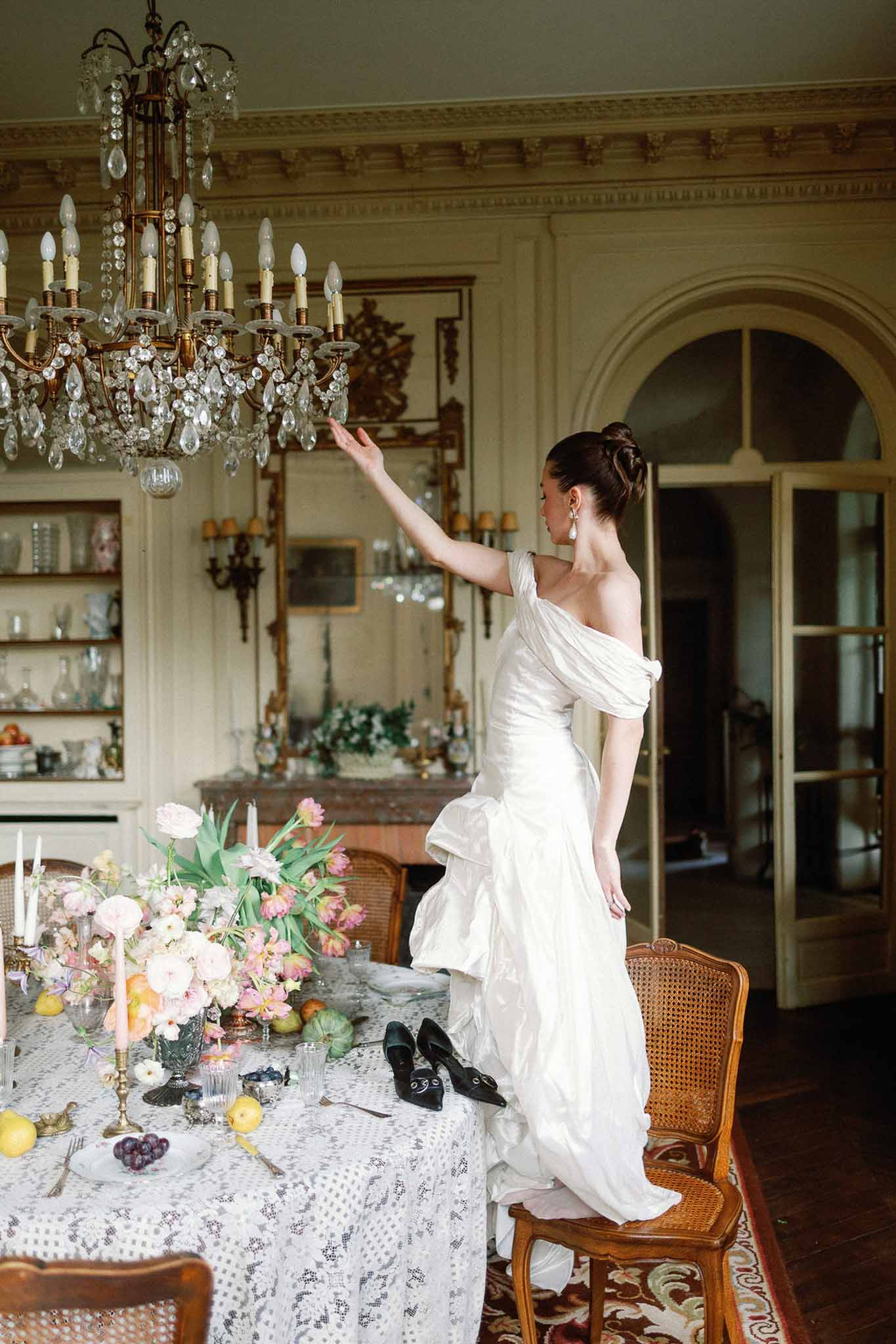 Bride standing on chair reaching toward chandelier beside pink tulip and peach ranunculus table styling
