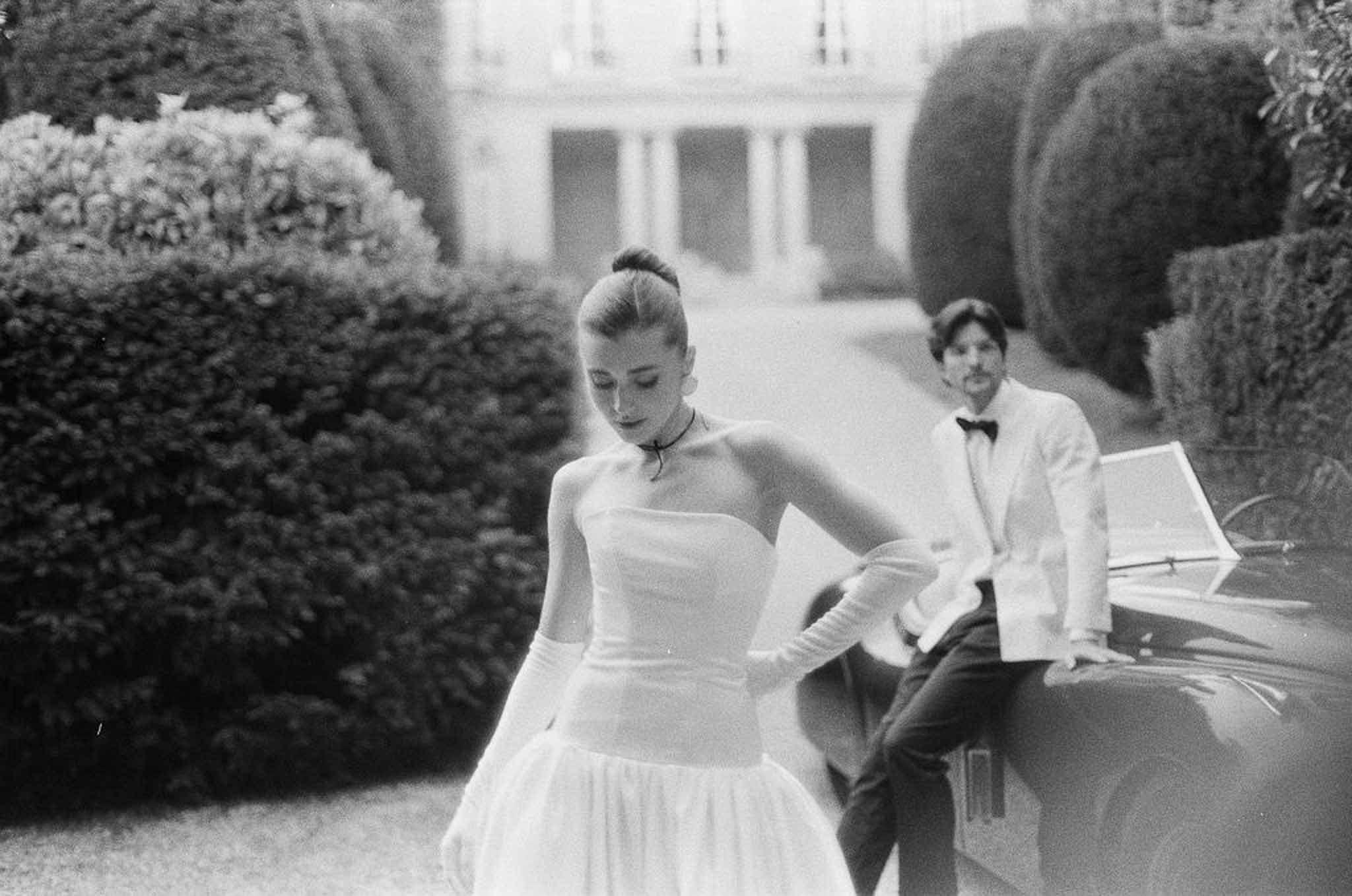 Black and white film portrait bride in ball gown with opera gloves groom leaning on vintage car at manor