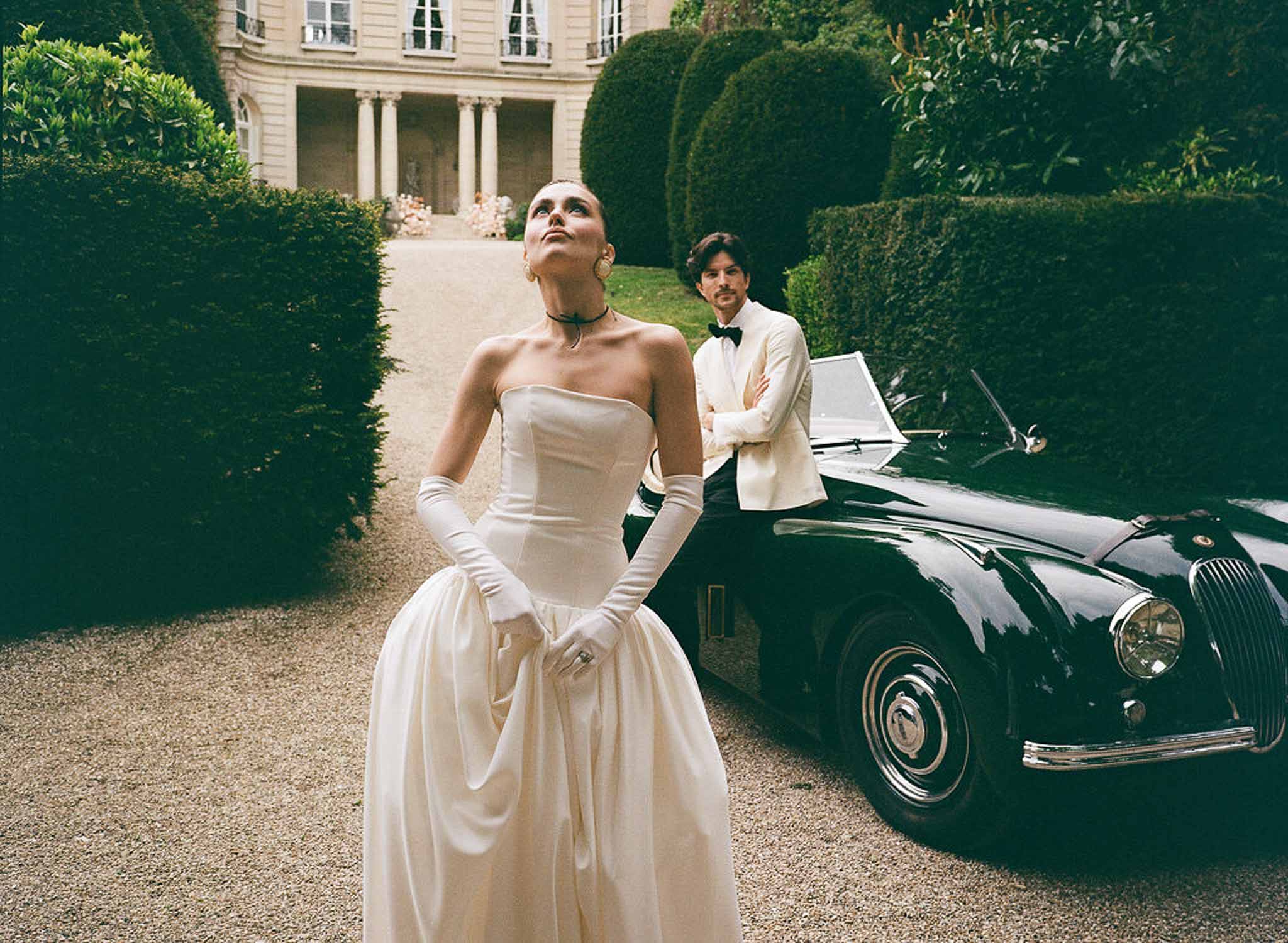 Bride in ballgown with opera gloves beside groom leaning on vintage Jaguar at columned manor