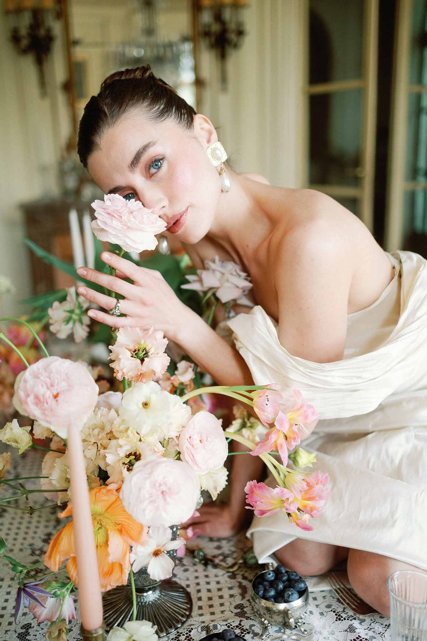 Bride in ivory strapless gown with statement earrings leaning close to blush rose and coral tulip centerpiece