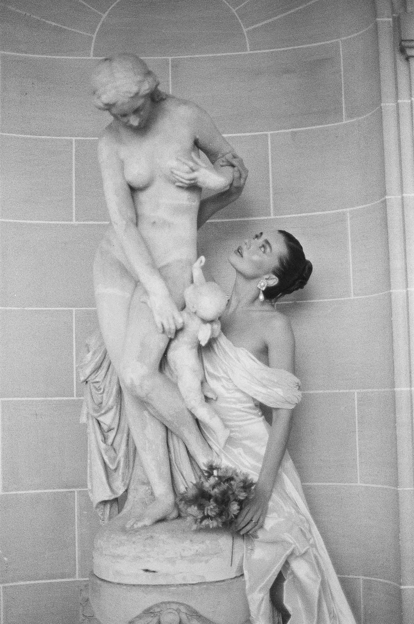 Black and white film portrait of bride with bouquet beside classical marble statue in stone niche