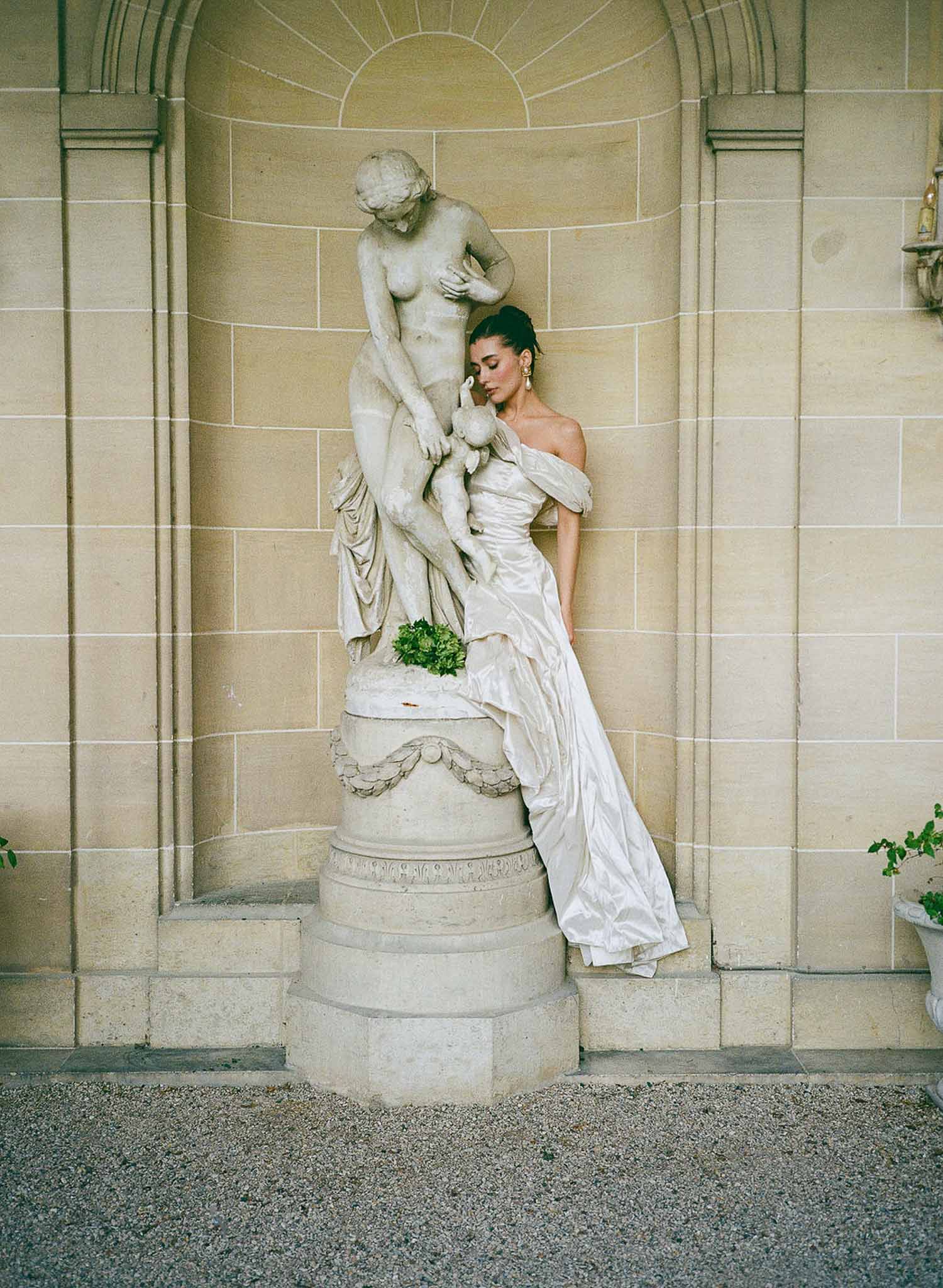 Bride in off-shoulder ivory satin gown with cascading train posed against neoclassical stone sculpture niche
