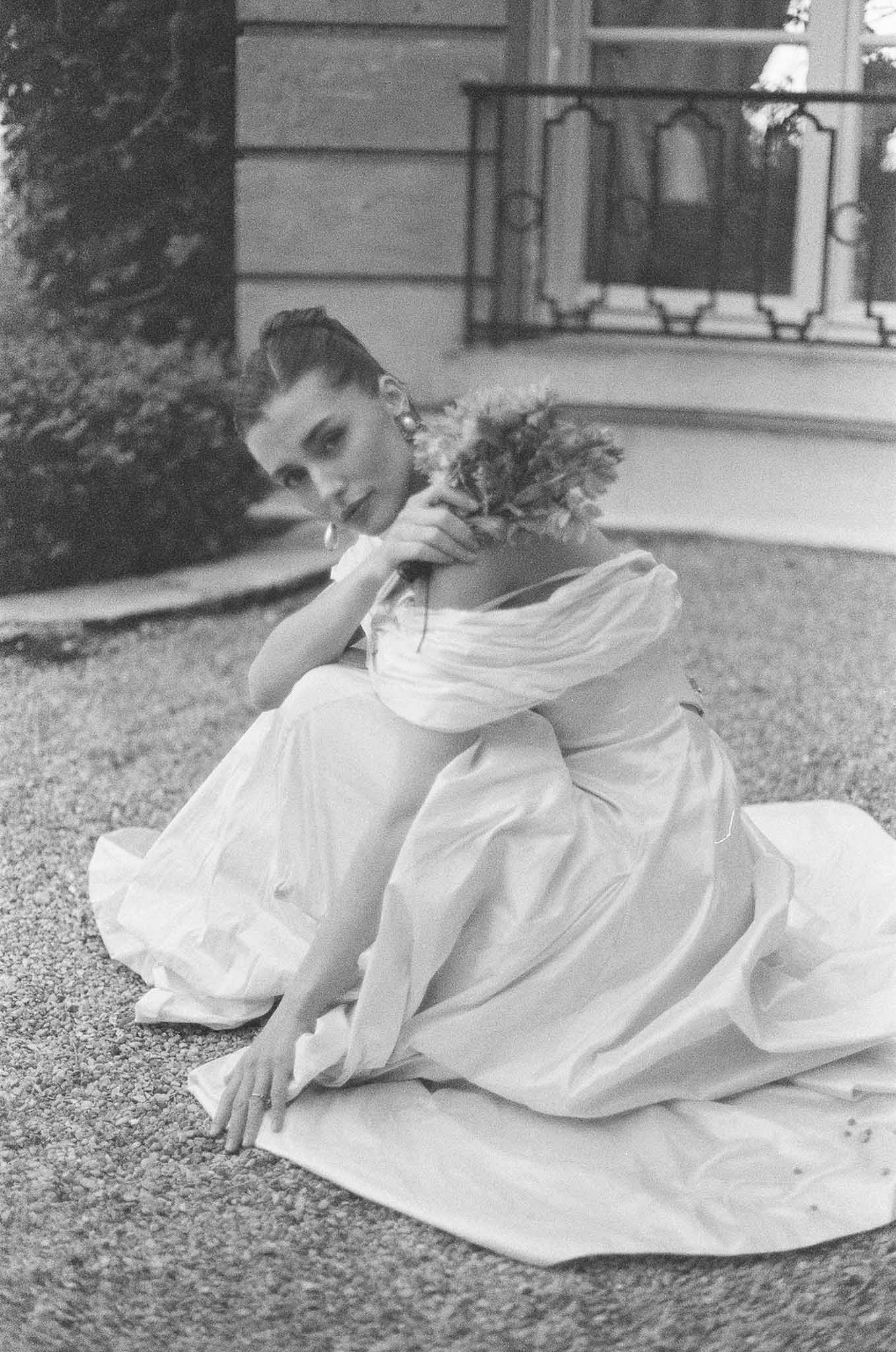 Bride in off-shoulder ballgown seated on gravel before chateau balcony looking at camera in B&W film
