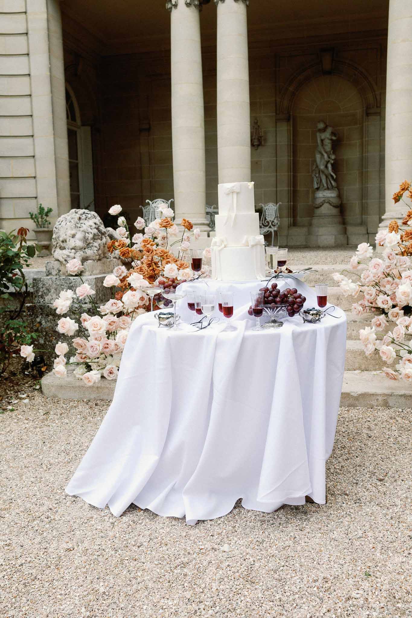 Four-tier white bow cake on draped table with cascading blush and burnt orange roses at chateau steps