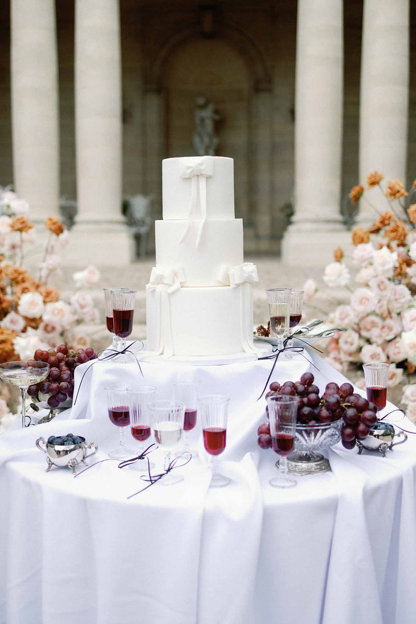 Four-tier ivory wedding cake with blush and terracotta roses, crystal glassware, and stone colonnade backdrop