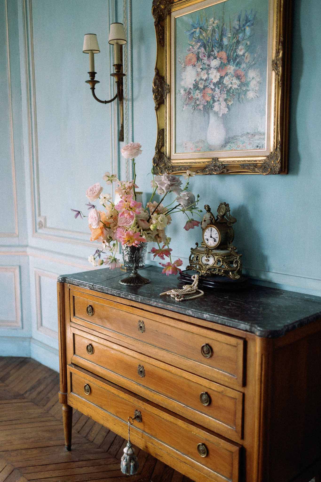 Floral arrangement of blush roses and coral cosmos on antique French chest with gilded clock and pearl necklace