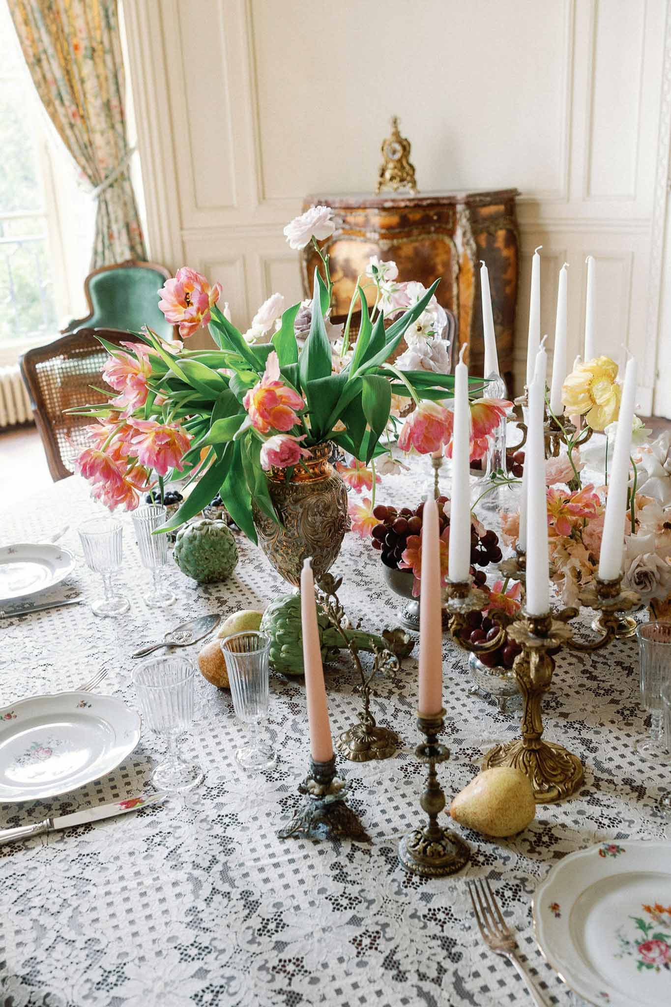 Maximalist tablescape with silver urn of pink tulips, brass candelabras, grapes, and floral porcelain plates