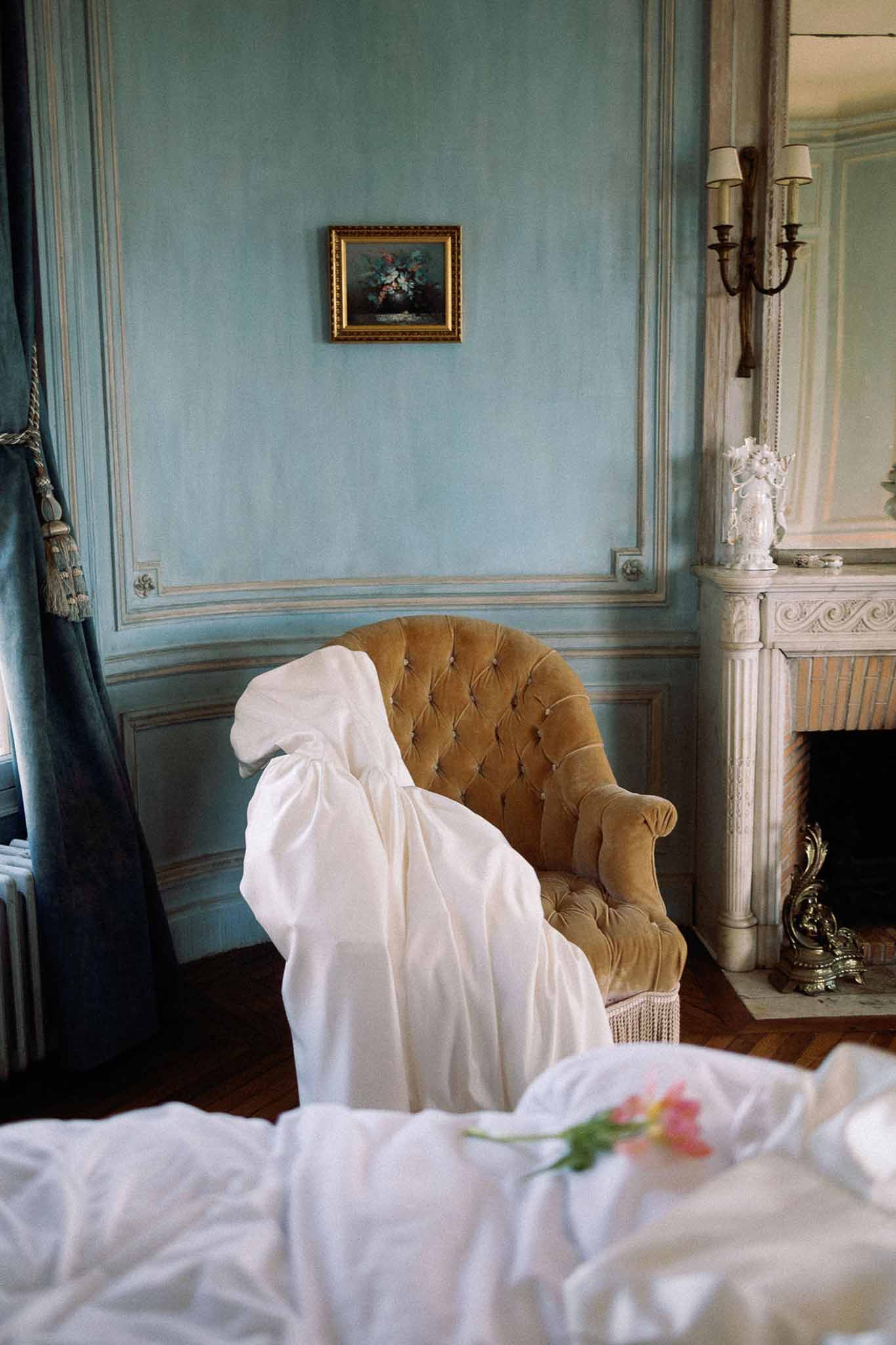 Ivory wedding dress draped on gold velvet chair in blue-panelled chateau bedroom with marble fireplace