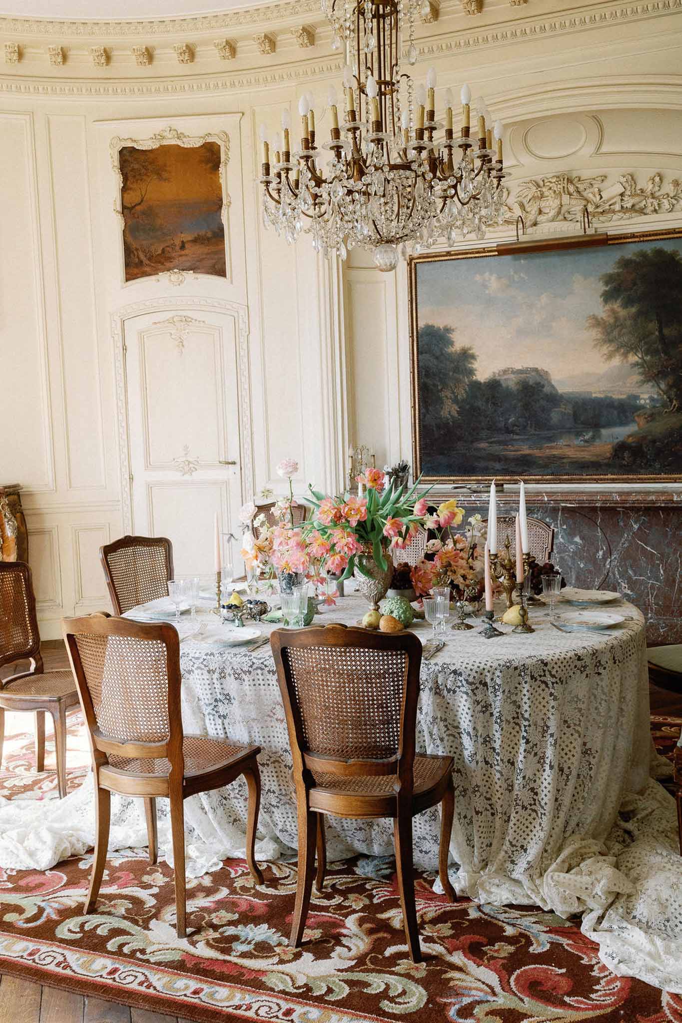 Oval reception table with white lace cloth, coral tulip centerpiece, taper candles, and crystal glassware in chateau dinin...